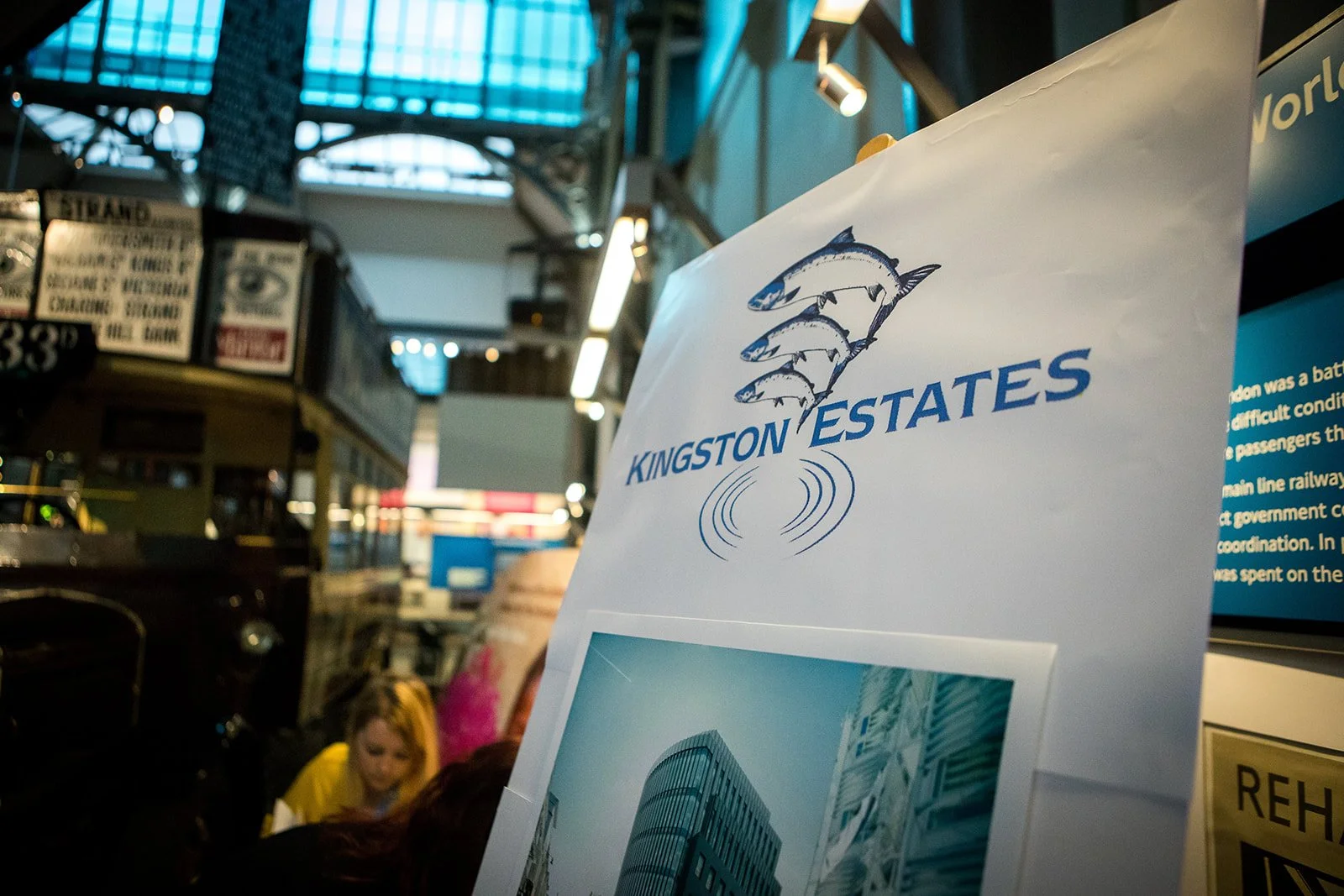A sign for Kingston Estates with a logo of three fish jumping above water, located in an indoor setting with large glass windows and a high ceiling, possibly a transportation hub or shopping area.