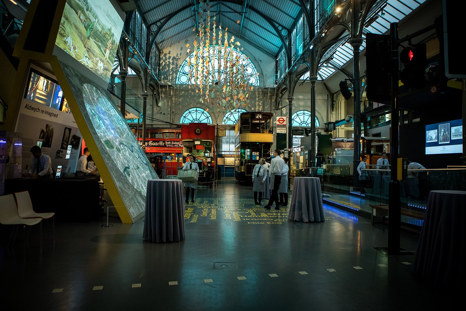 Interior of a train station or transport hub with vintage-style trains, informational displays, and modern lighting fixtures inside a large glass-roofed building.