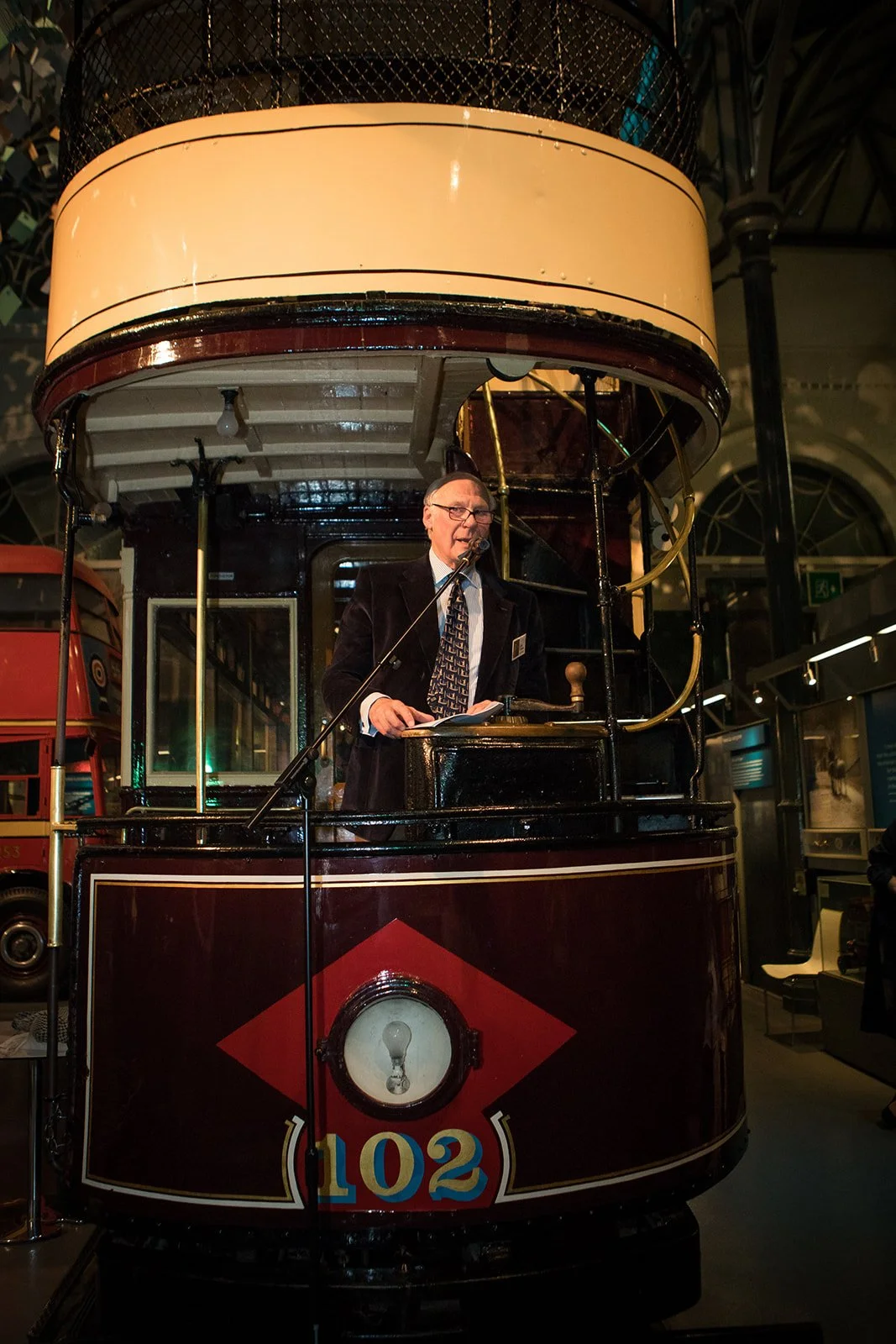 A man wearing glasses and a black suit with a patterned tie, standing inside a vintage tram or cable car, speaking into a microphone.