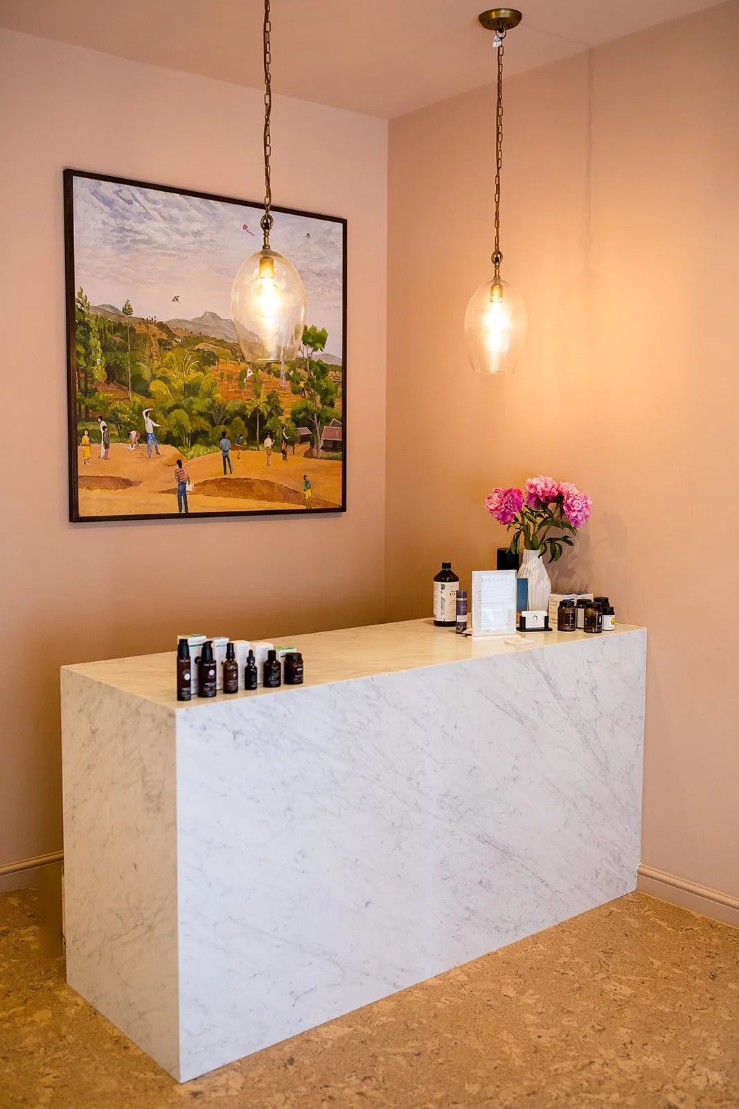 A reception desk with bottles and a sign, pink flowers in a white vase, wall art showing a landscape scene with people, and two hanging light fixtures in a room with beige walls and cork flooring.