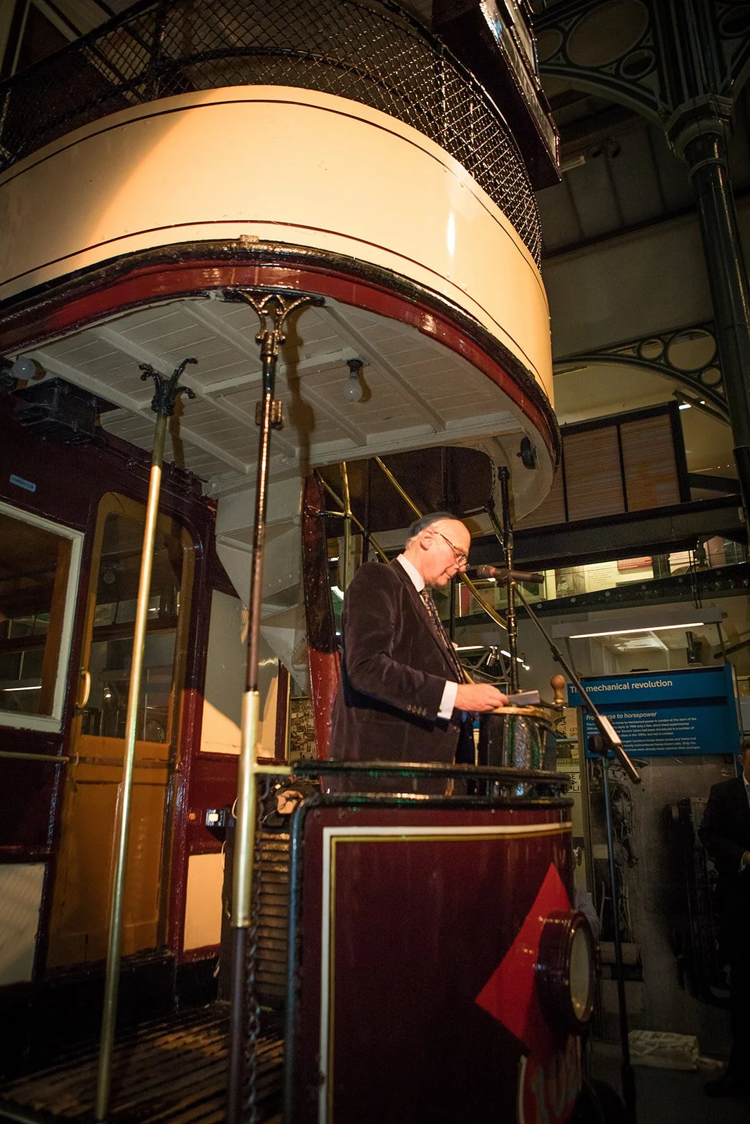 A man in a suit and glasses reading from a paper at a microphone, standing on a vintage streetcar inside a museum or exhibition.