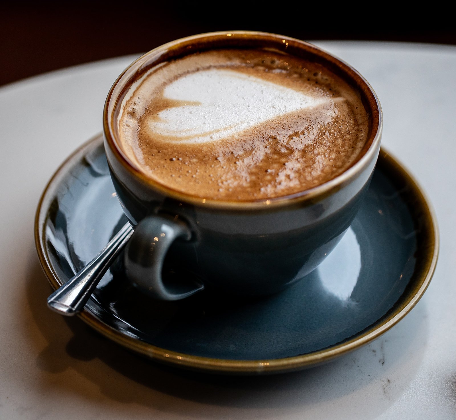 A cup of coffee with foam art served in a dark ceramic mug on matching saucer, with a teaspoon resting on the side.