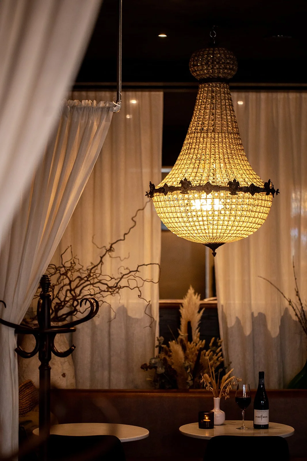 Elegant chandelier hanging from ceiling above a small round table with a bottle of wine, glass of red wine, and decorative dried plants in a white vase. Soft lighting and curtains create a cozy, upscale ambiance.