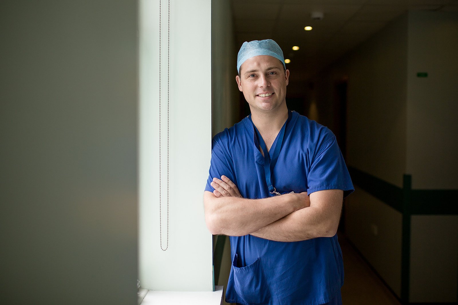 A smiling male medical professional wearing blue scrubs and a disposable cap, standing with arms crossed near a window in a hospital corridor.