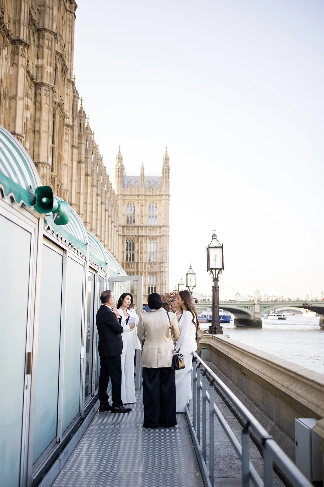 A group of four businesspeople, two men and two women, are having a discussion on a riverside terrace with a view of the Palace of Westminster in London. The group is dressed in formal business attire, and the setting features historic street lamps a