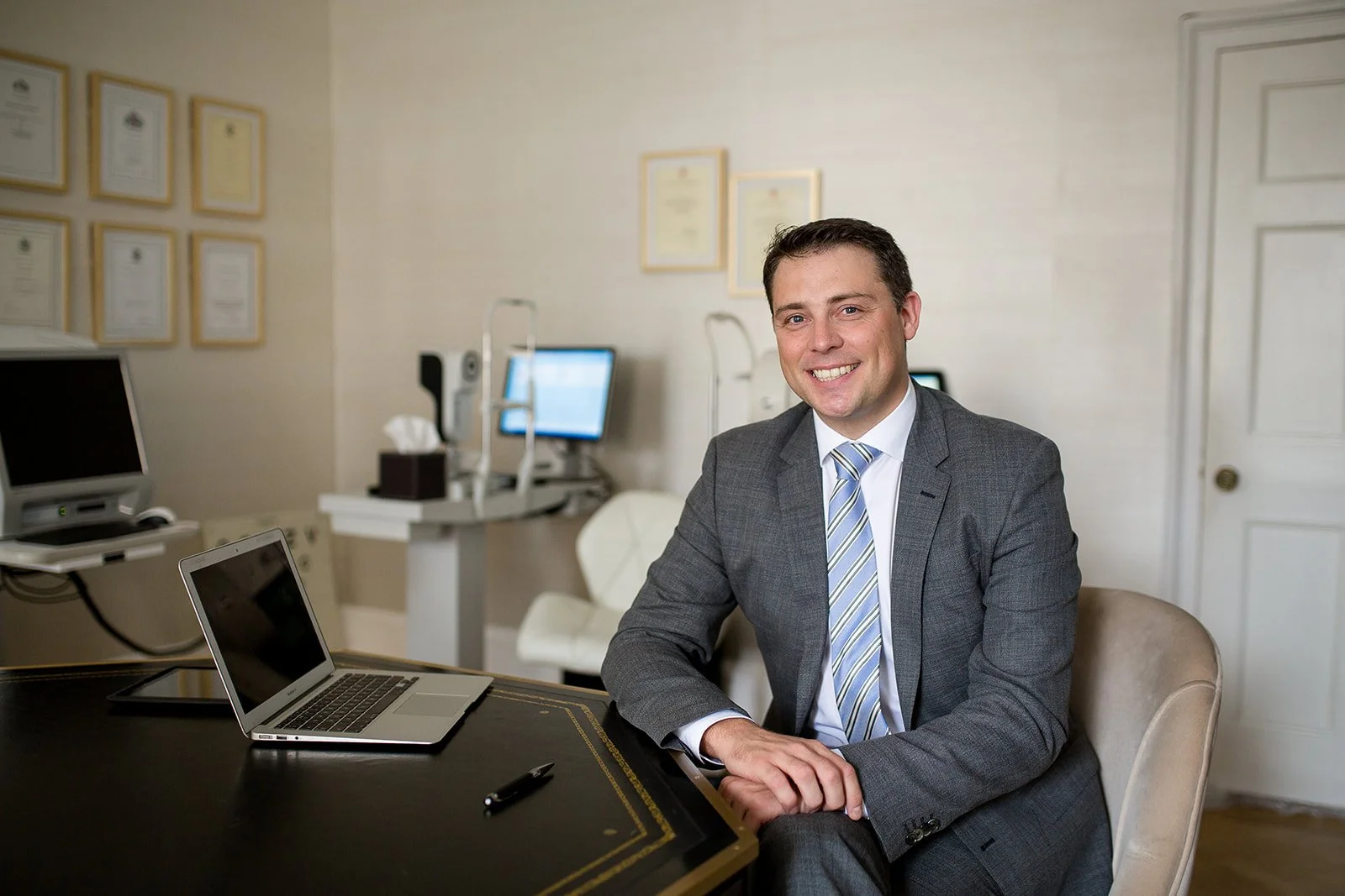 A man in a gray suit smiling and sitting at a desk with a laptop, in an office setting with computer monitors and framed certificates on the wall.