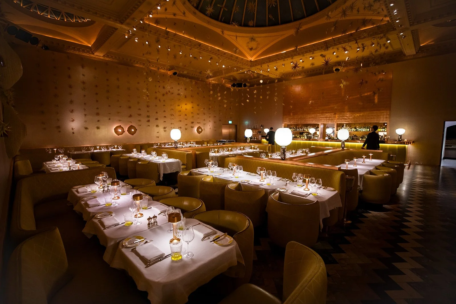 Elegant restaurant dining area with neatly set tables, ambient lighting, and a bar in the background decorated with star-shaped ornaments on the ceiling and walls.
