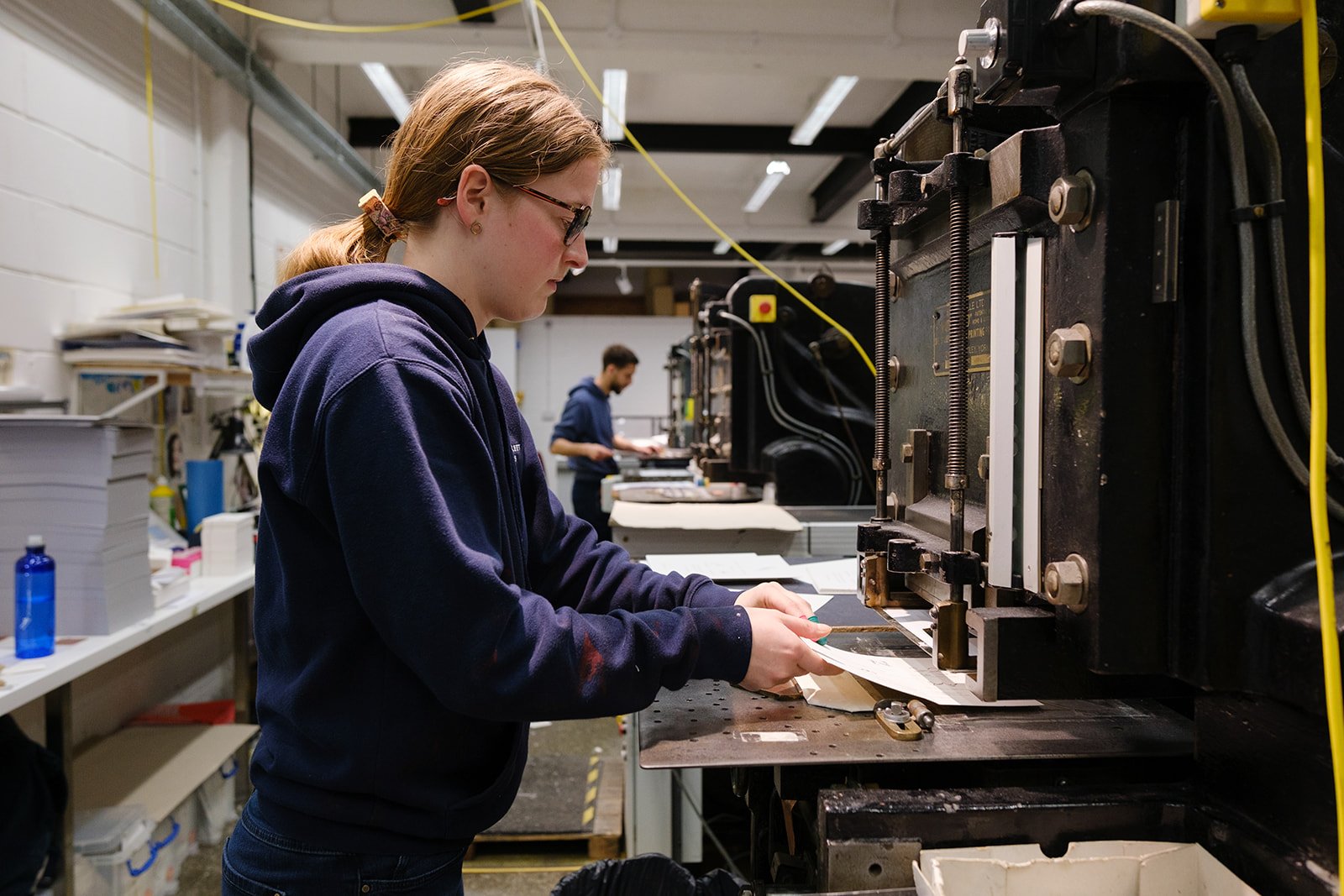 A young woman with glasses working on a printing machine in a workshop, with another person in the background also working at a station.