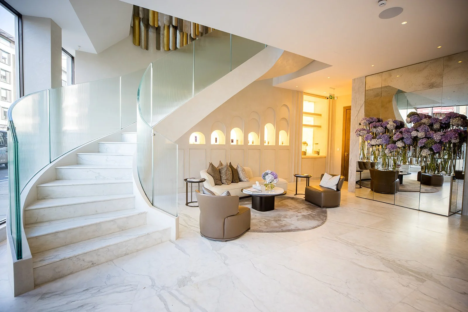 Elegant hotel lobby with a white marble spiral staircase, a cozy sitting area with a sofa, armchairs, and a coffee table decorated with purple flowers, and large mirrors reflecting purple hydrangeas in vases. Warm lighting creates a welcoming atmosph