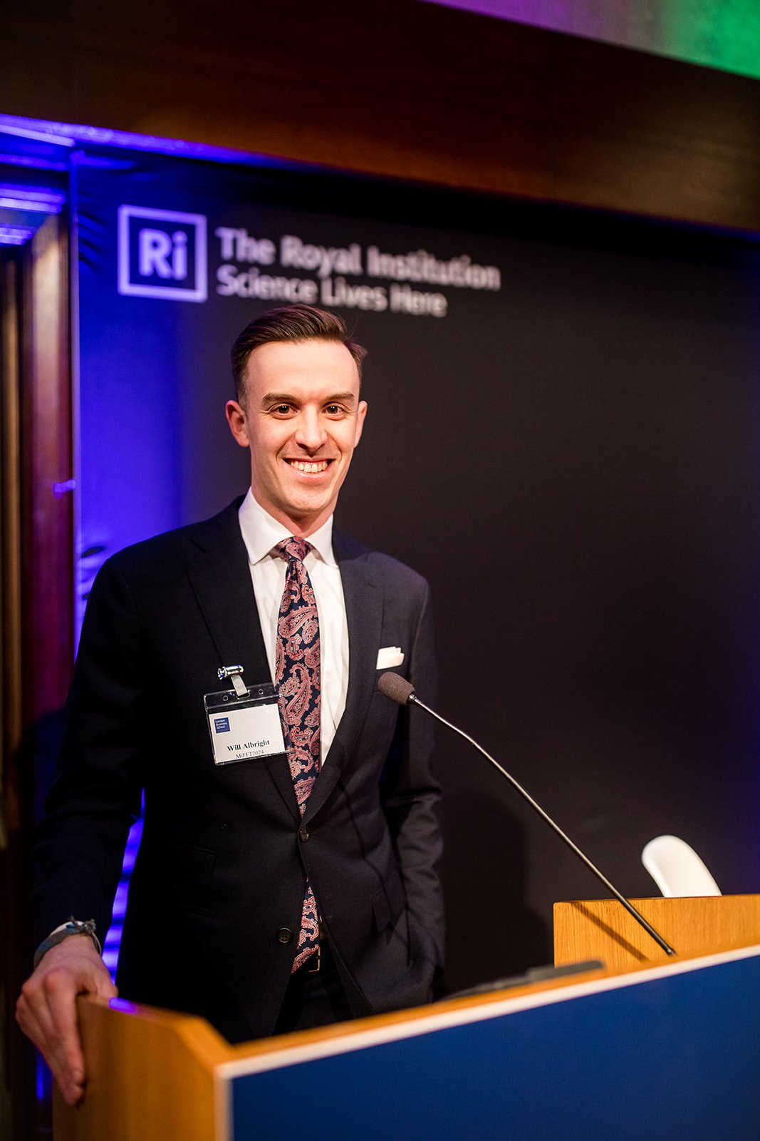 A man in a black suit and colorful tie standing at a podium with a microphone, smiling, in front of a sign that reads "The Royal Institution Science Lives Here" with a logo that says Ri.