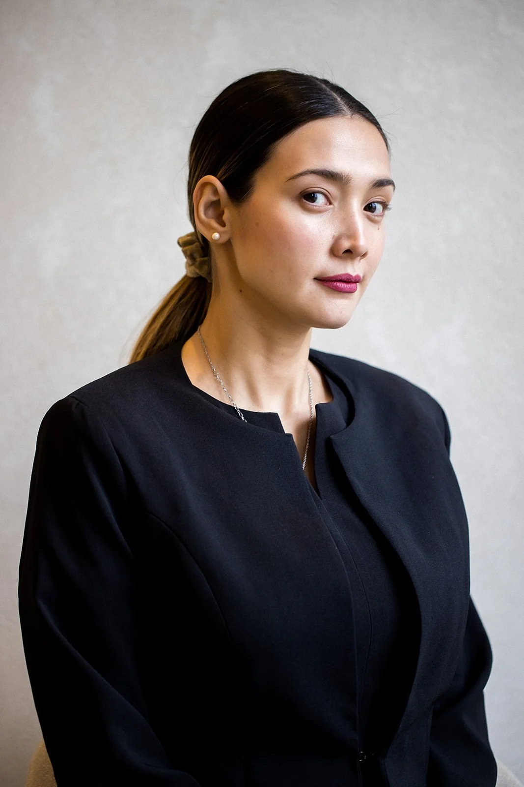 A woman with dark hair tied back, wearing a black top, pearl earrings, a necklace, and makeup, standing against a plain light-colored background.