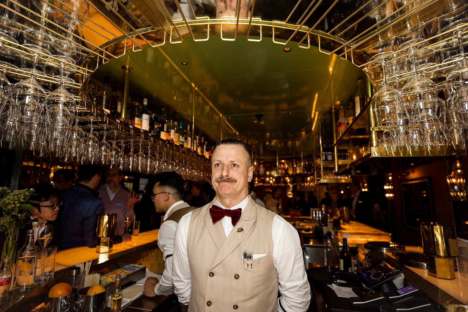 A bartender with a mustache and beige vest, wearing a red bow tie, stands behind a bar with hanging glasses and bottles, in a busy, warmly lit bar or restaurant.