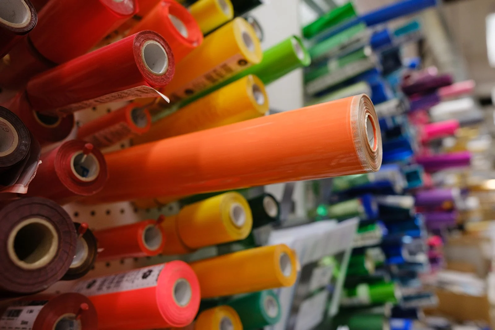 Colorful rolls of vinyl or wrapping paper stored on a shelf in various shades including red, orange, yellow, green, blue, and purple, in a store.
