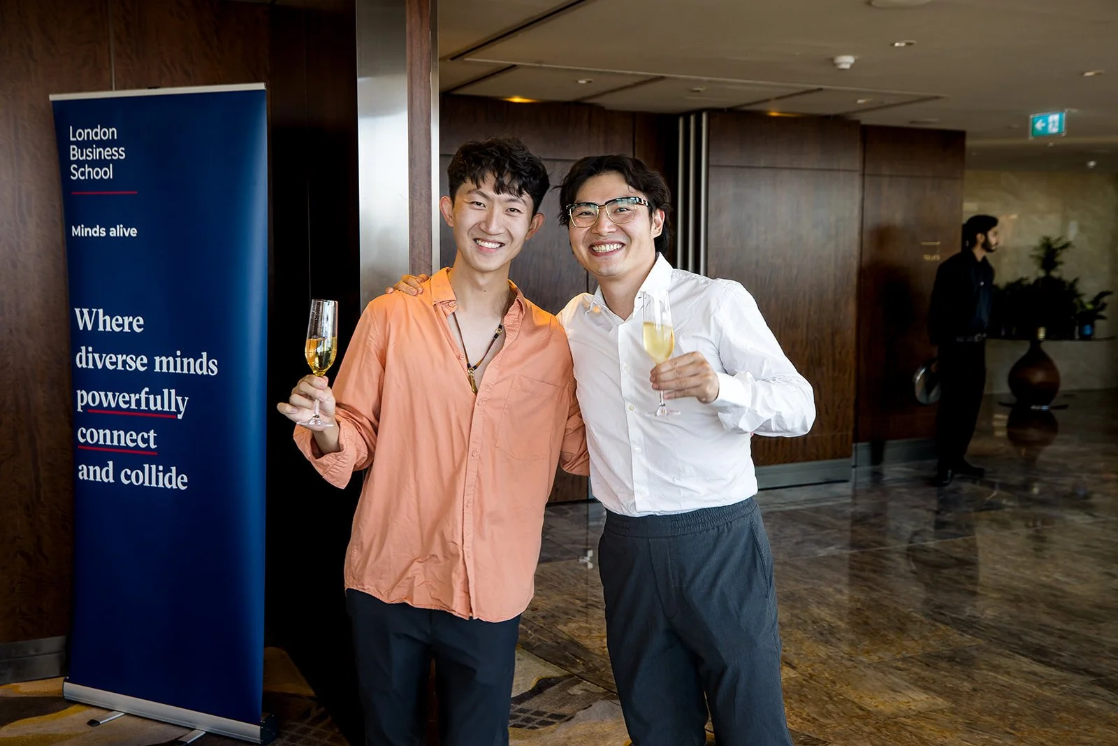 Two men smiling at the camera while holding glasses of champagne at an event. A London Business School banner is visible behind them.