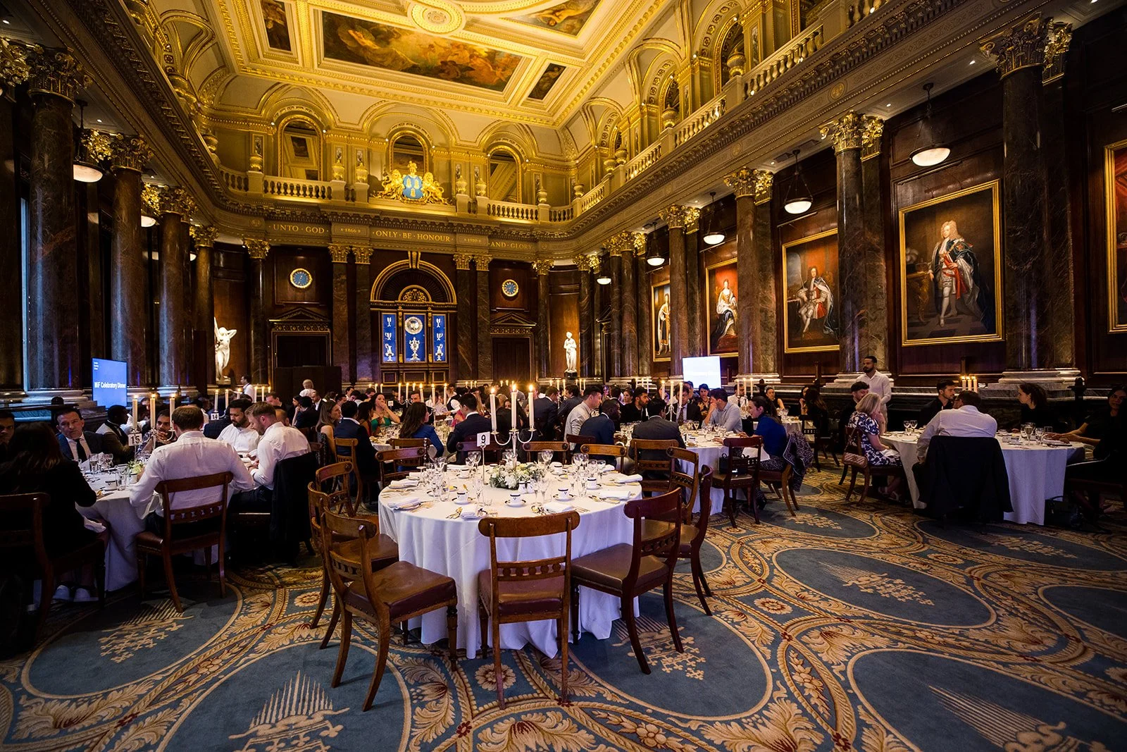 A grand banquet hall with ornate gold and dark wood decor, large paintings of historical figures on the walls, and multiple round tables set with white tablecloths, candles, and fine china for a formal dinner event.