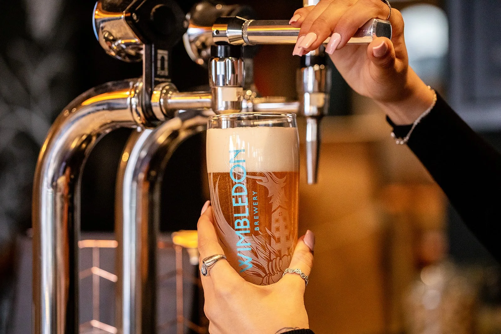 A person is filling a glass of beer from a tap, with the glass labeled "WiMBLEMDED ON BREWERY." The person's hand is adorned with rings and a bracelet, and they have manicured nails. The background is blurry with warm lighting.