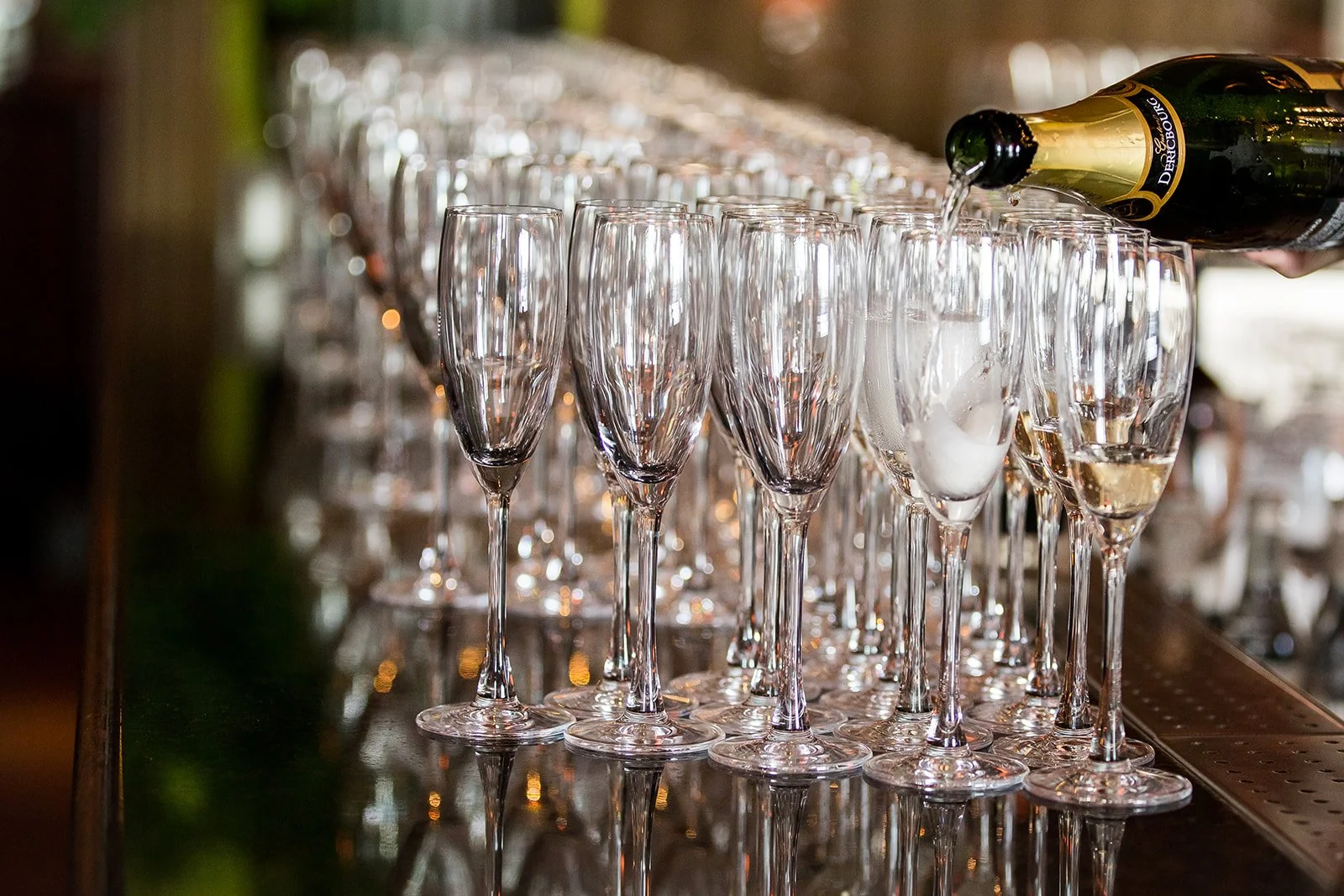 A row of empty champagne flutes on a bar counter with someone pouring a bottle of champagne into one glass.