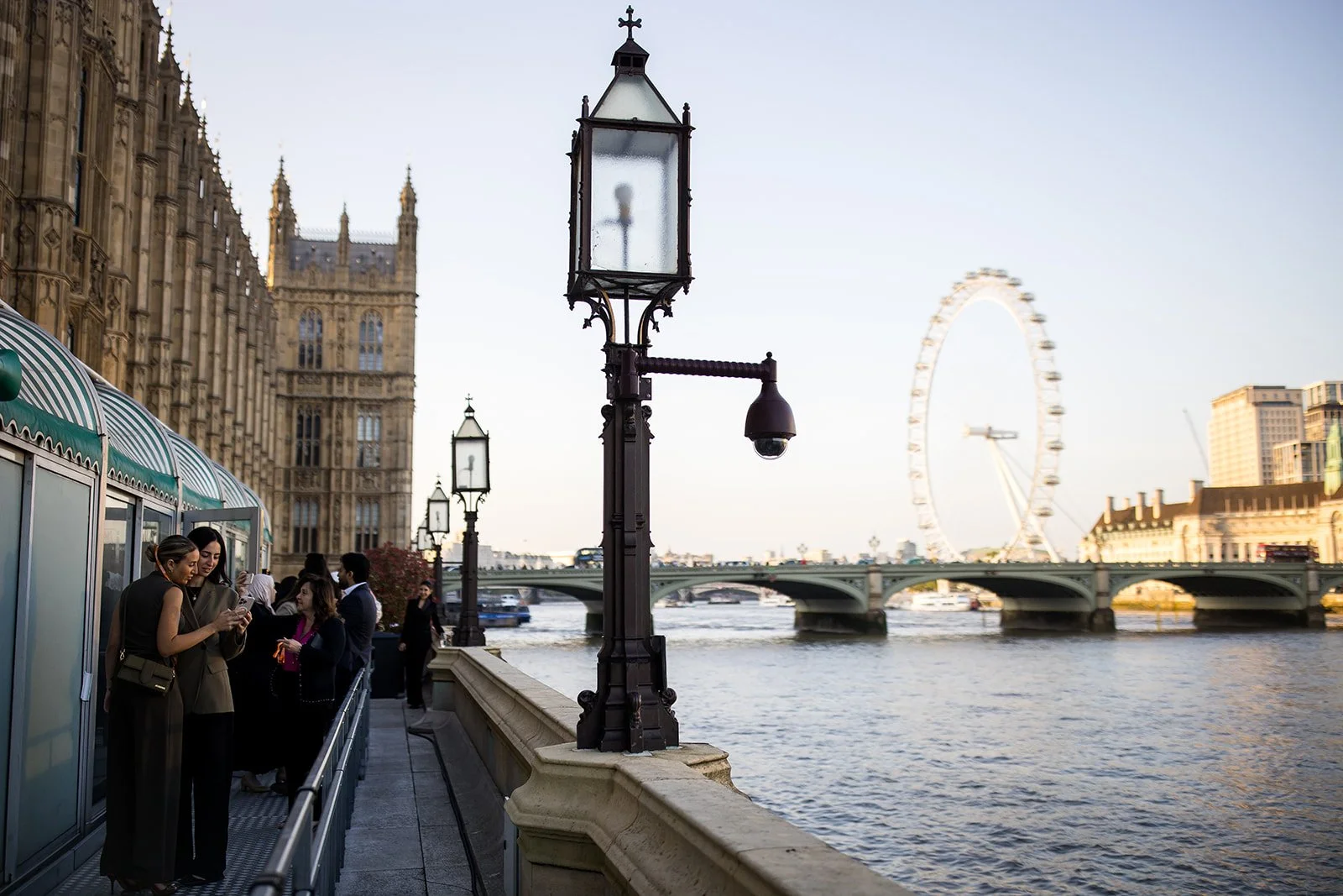 People standing on a riverside walkway in London, with a view of the River Thames, Westminster Bridge, the London Eye, and historic buildings.