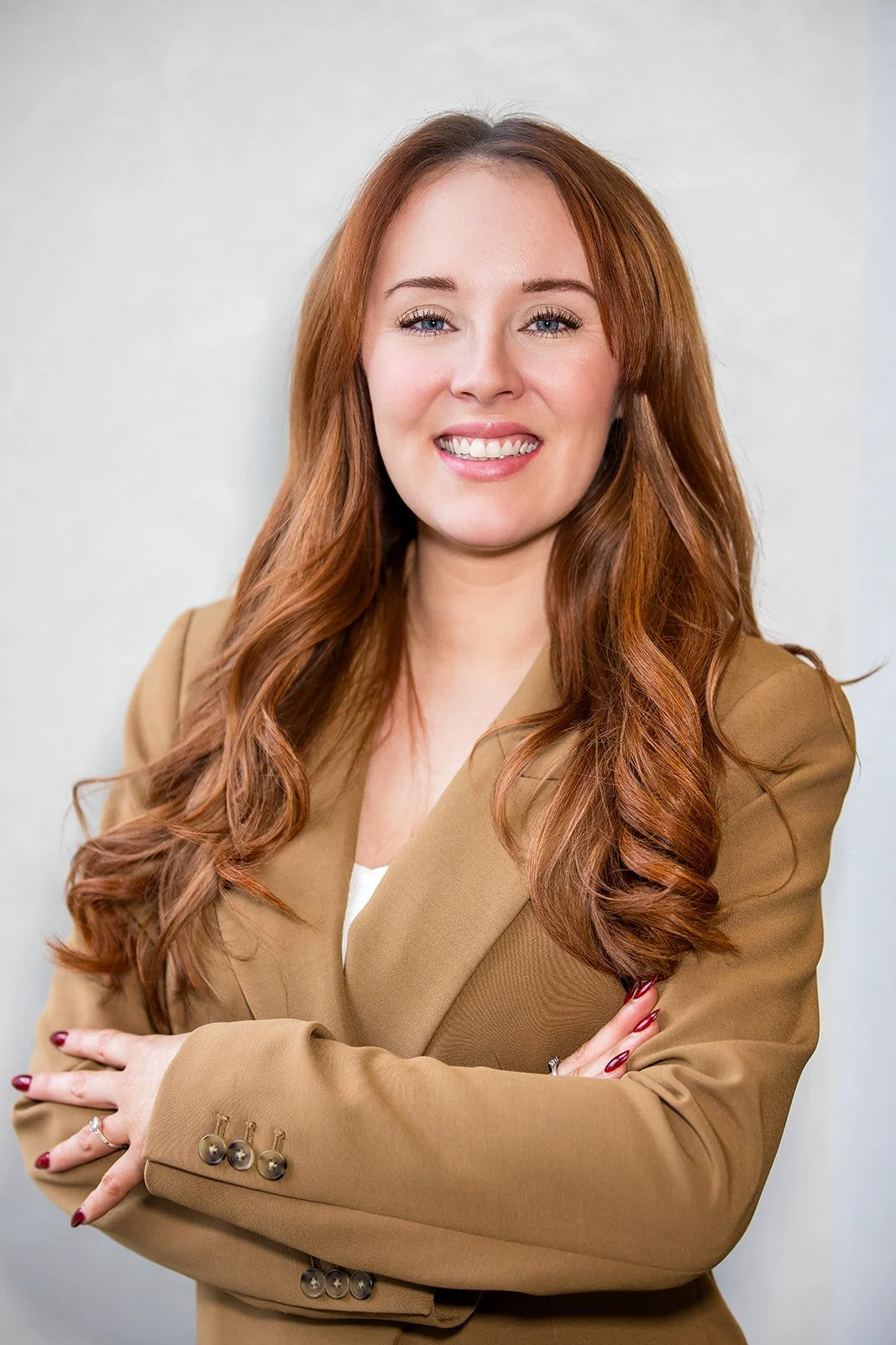 A smiling woman with long red hair in a tan blazer, crossing her arms. She has light skin, blue eyes, and is wearing makeup and red nail polish.