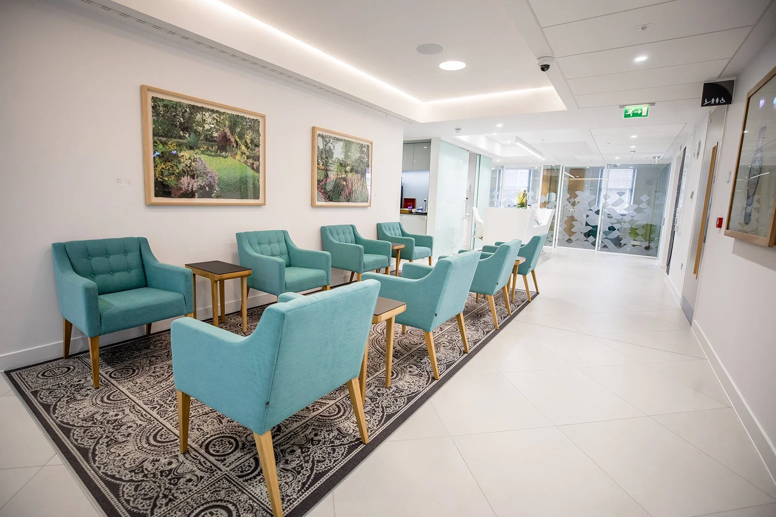 A modern waiting area with light blue chairs, small tables, framed artwork, and a patterned rug on a white tiled floor, with glass doors and windows in the background.