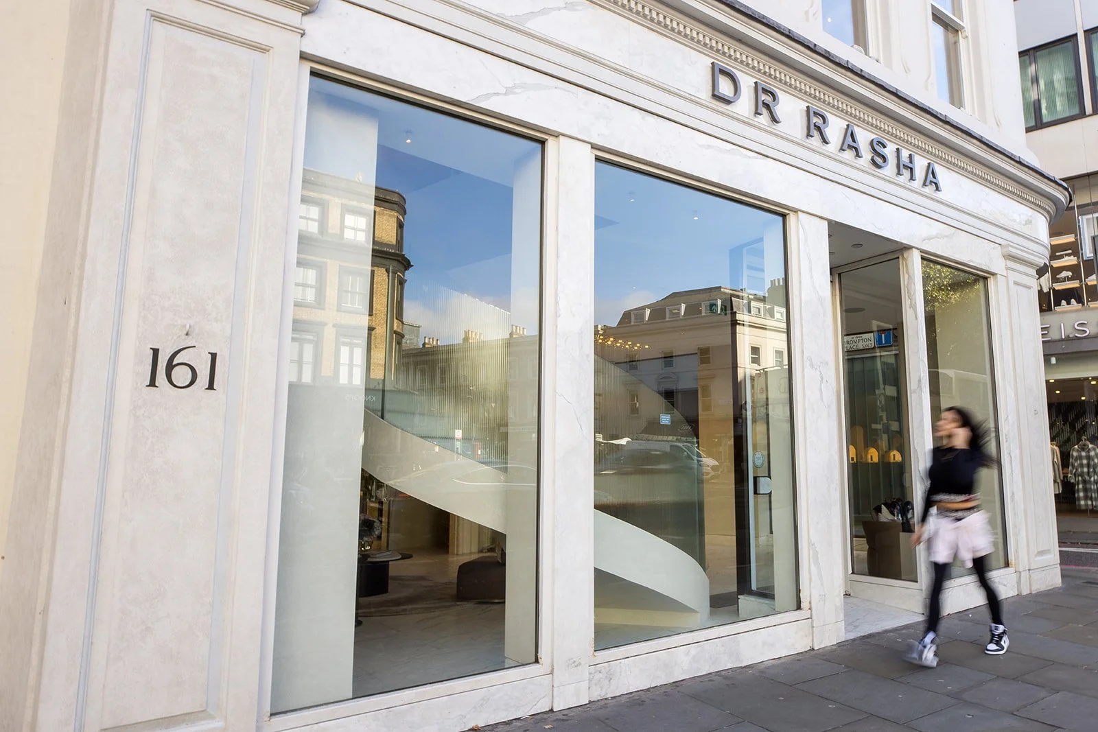 The storefront of D.R. Rasha, with large glass windows and a white marble facade. The address number is 161. A woman with long dark hair, wearing a black top, pink jacket tied around her waist, black leggings, and athletic shoes is walking past the s