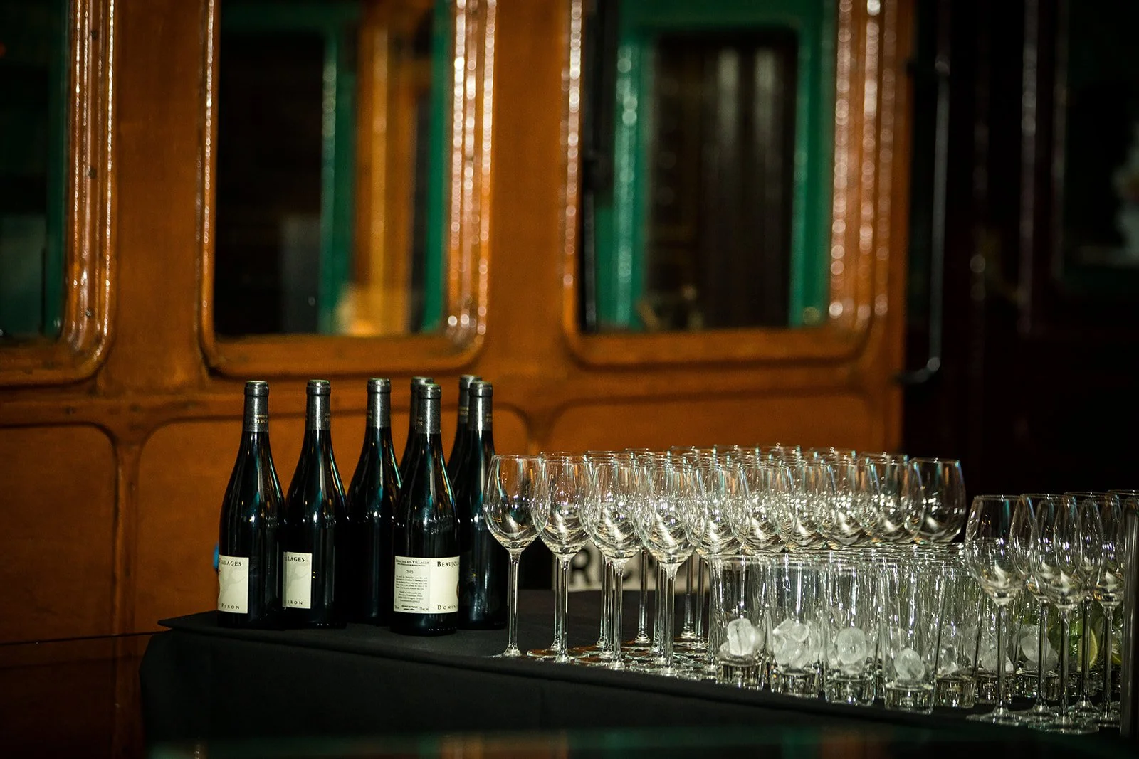 Bottles of wine and empty wine glasses on a table set for an event or celebration, with a wooden cabinet with glass doors in the background.
