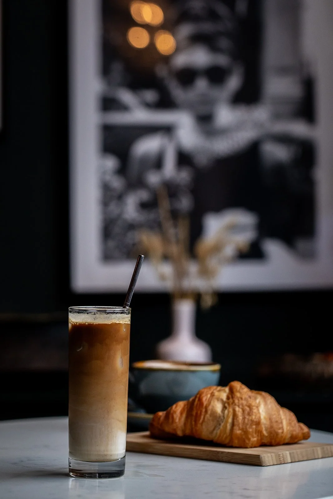 A glass of iced coffee with a straw, a croissant on a wooden cutting board, and a blue bowl in the background. There is a blurred framed artwork on the wall behind.