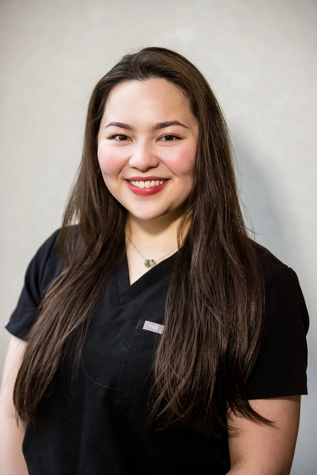 Portrait of a young woman with long brown hair, smiling, wearing a black medical uniform with a small silver tag that says 'FIGS'.