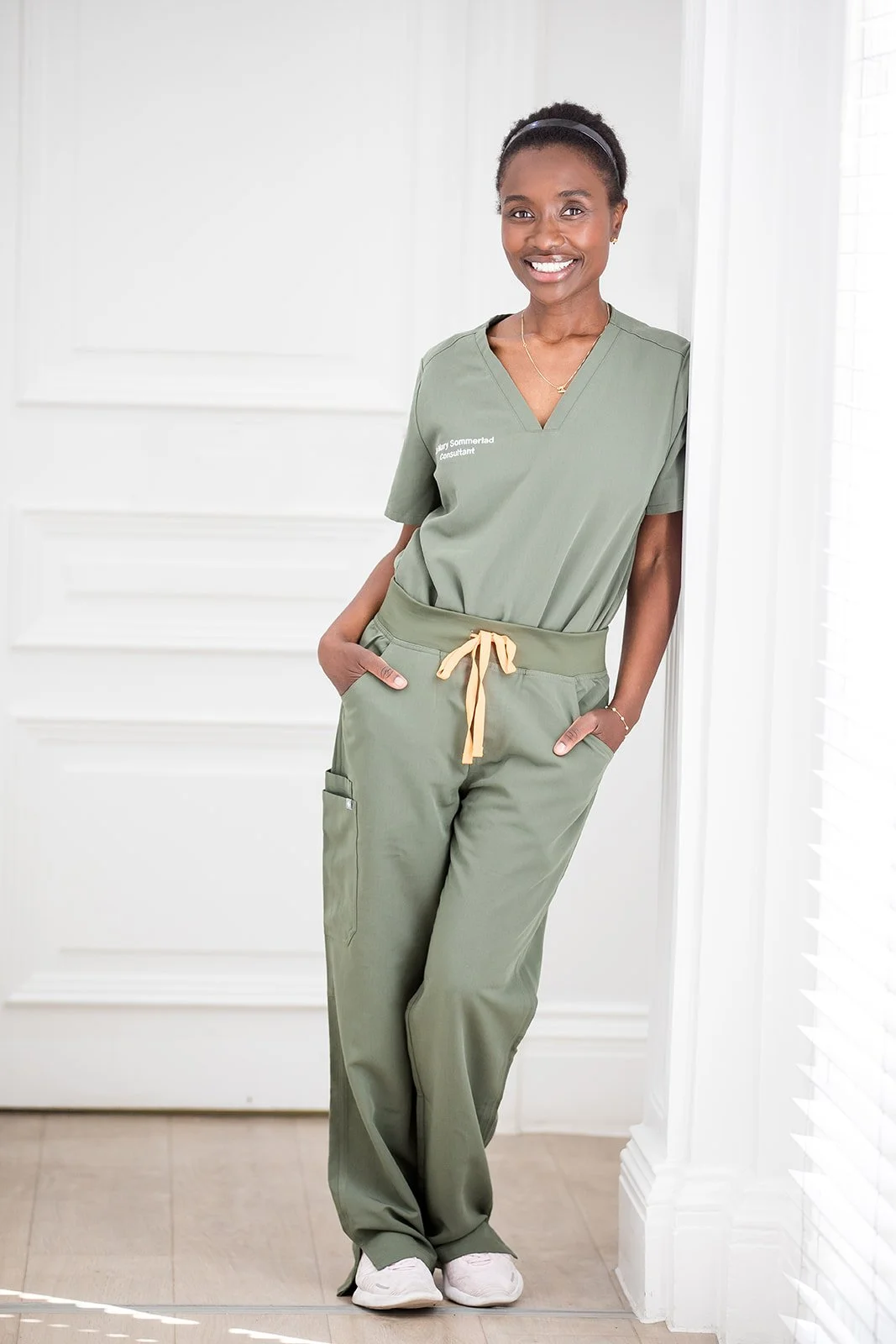 A smiling woman standing indoors, wearing light green medical scrubs, with her hands in her pockets and leaning against a white wall.