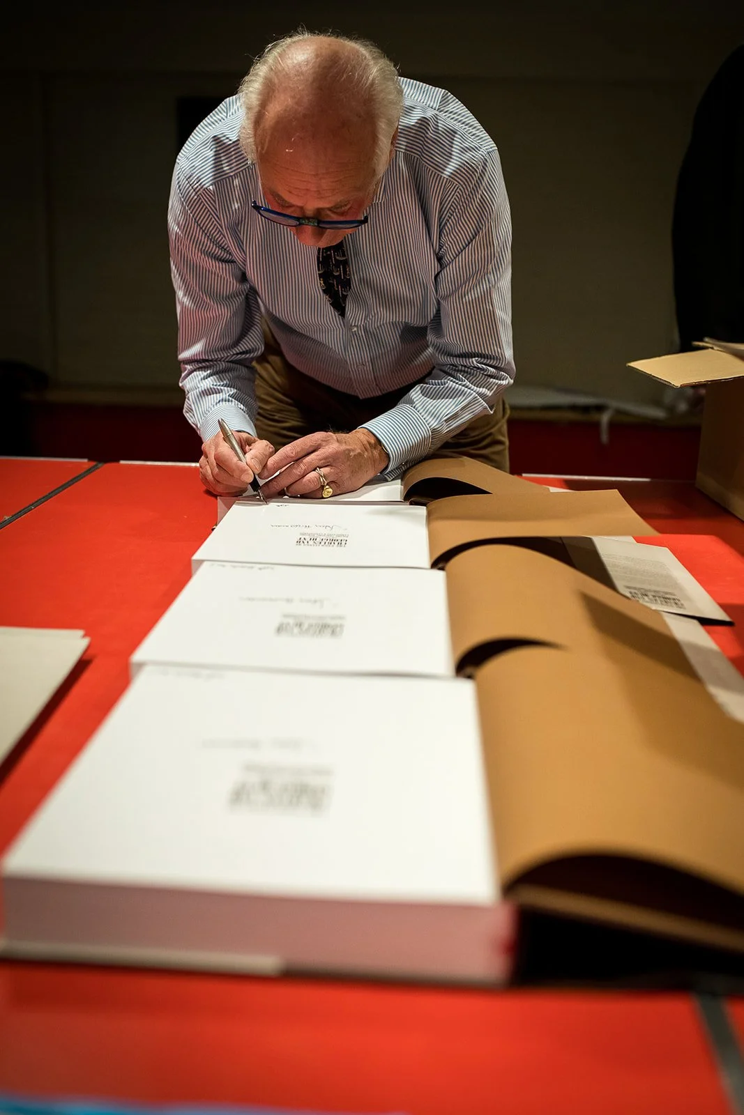 An elderly man with gray hair, wearing glasses and a striped shirt, signs a book on a red table, with several copies of the book laid out in front of him.