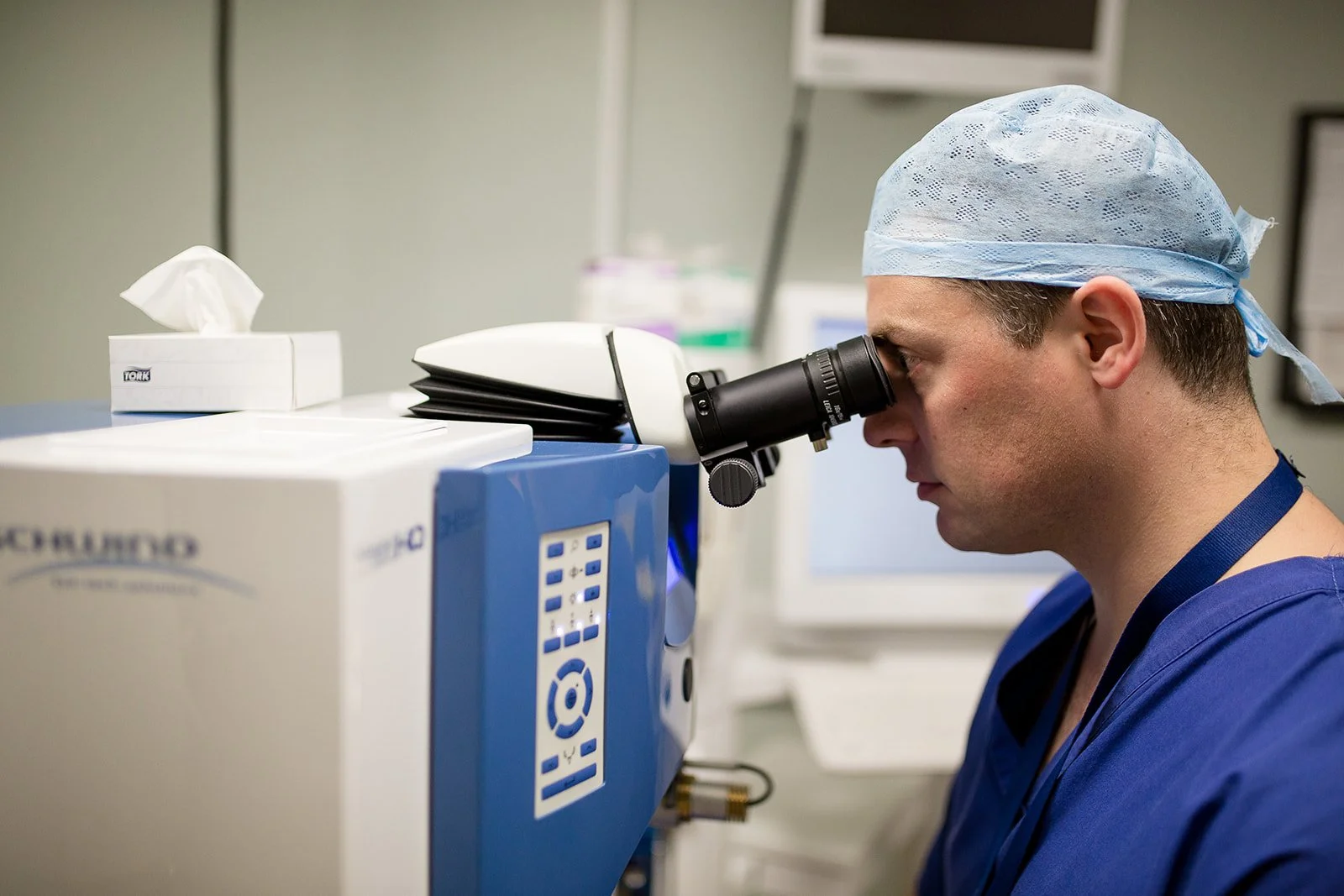 A healthcare professional wearing scrubs and a hair cap examines a sample through a microscope in a medical lab.