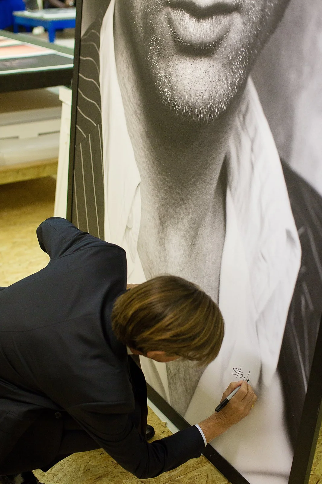 A man in a suit is signing a large black-and-white poster of a person’s face, focusing on the beard and lips, in an indoor setting.