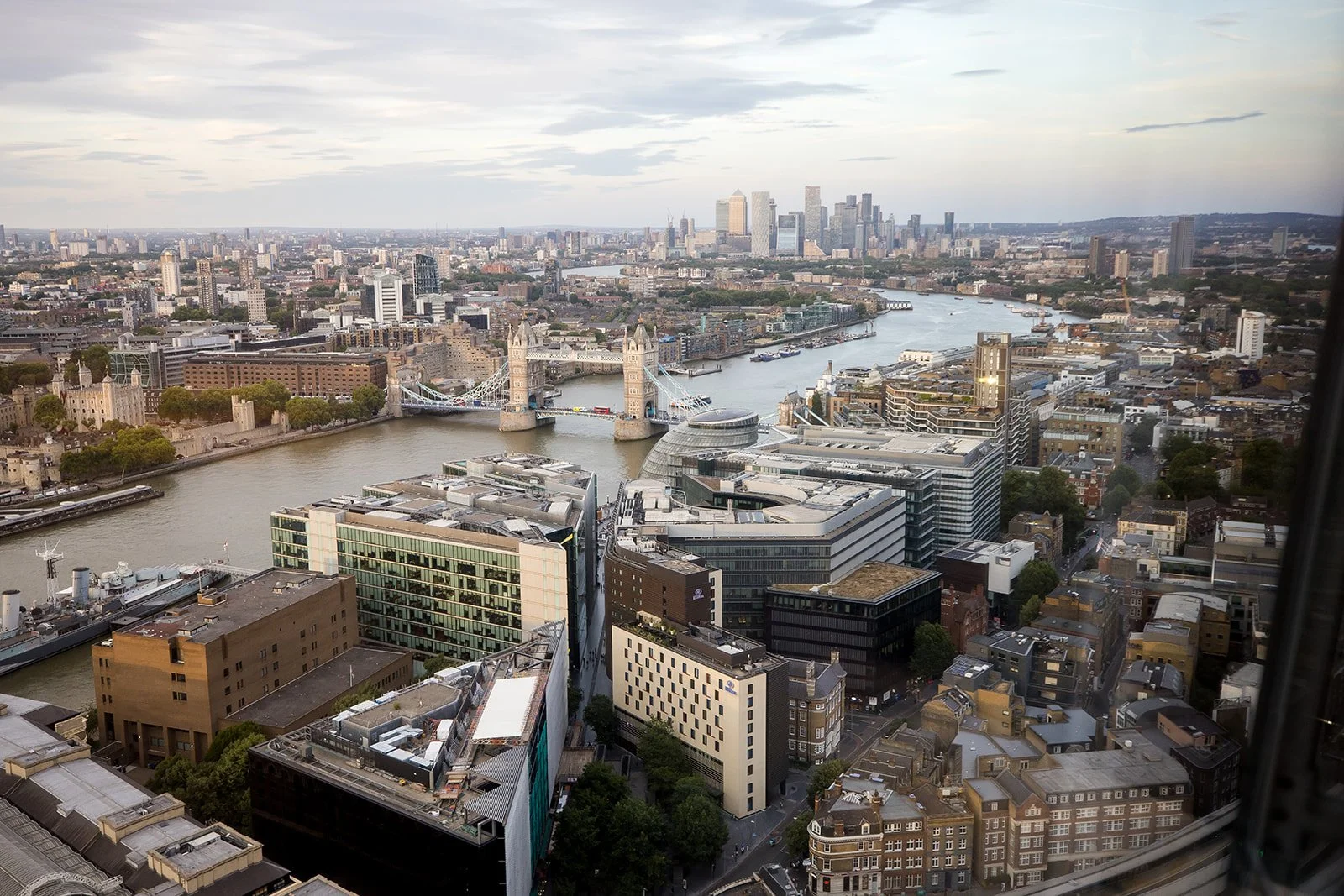 A high-angle view of London cityscape featuring the Tower Bridge crossing the River Thames, with modern and historic buildings, and a cluster of skyscrapers in the background.