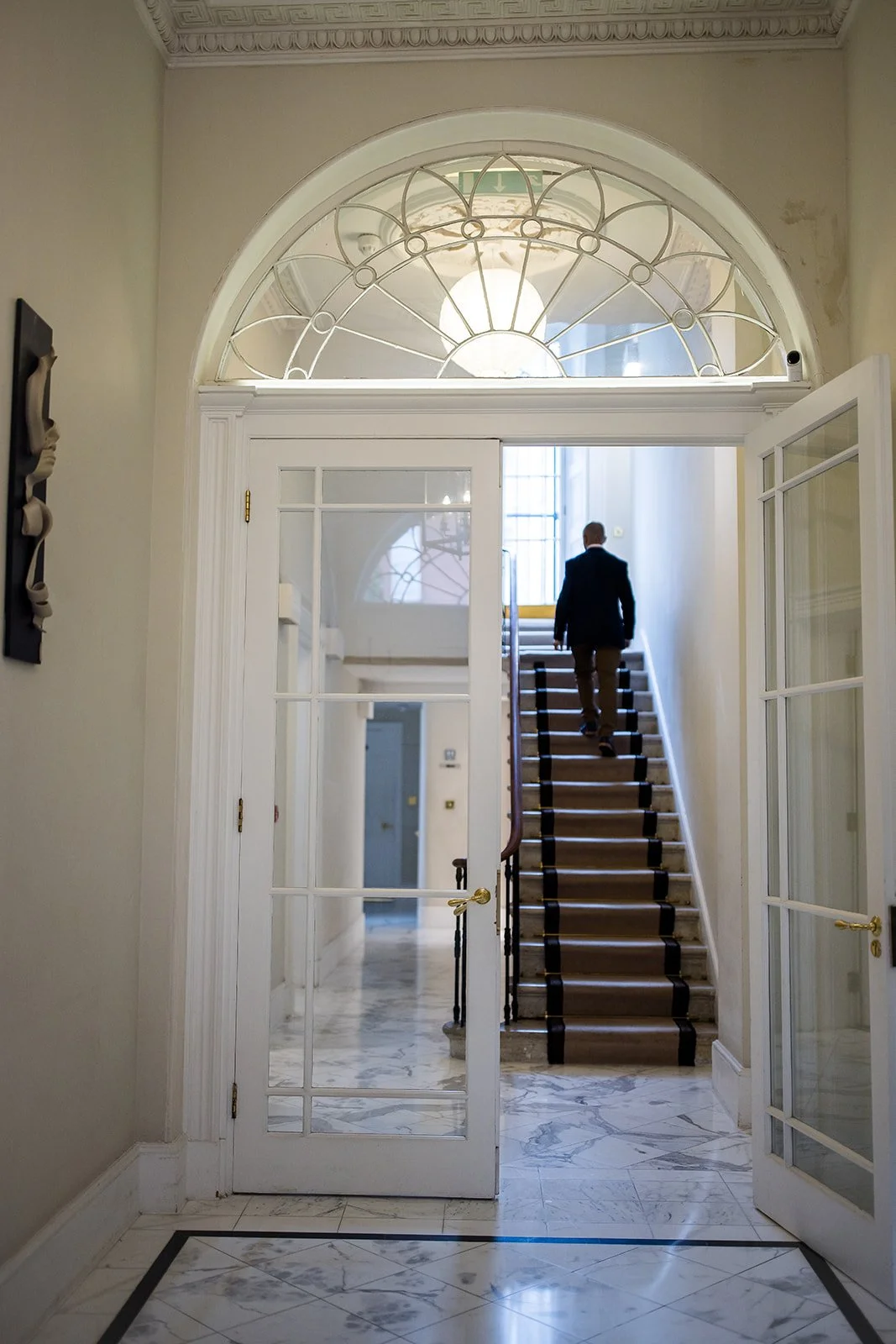 Interior view of a building entrance with a glass door, marble floor, staircase, and a man in a suit walking up the stairs.