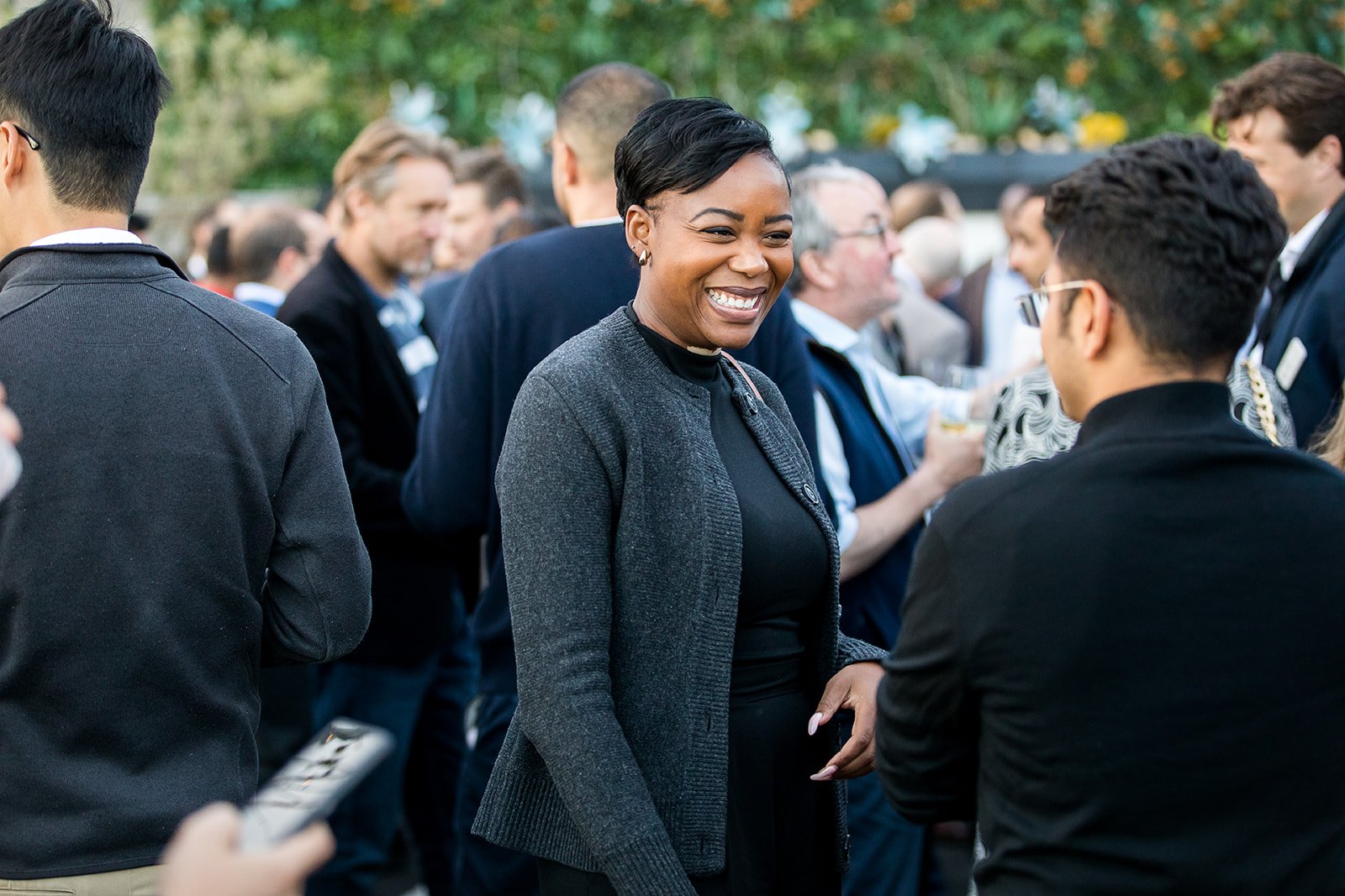 A group of people at an outdoor social event, with a woman smiling and talking to a man in the center, background with trees and blurred crowd.