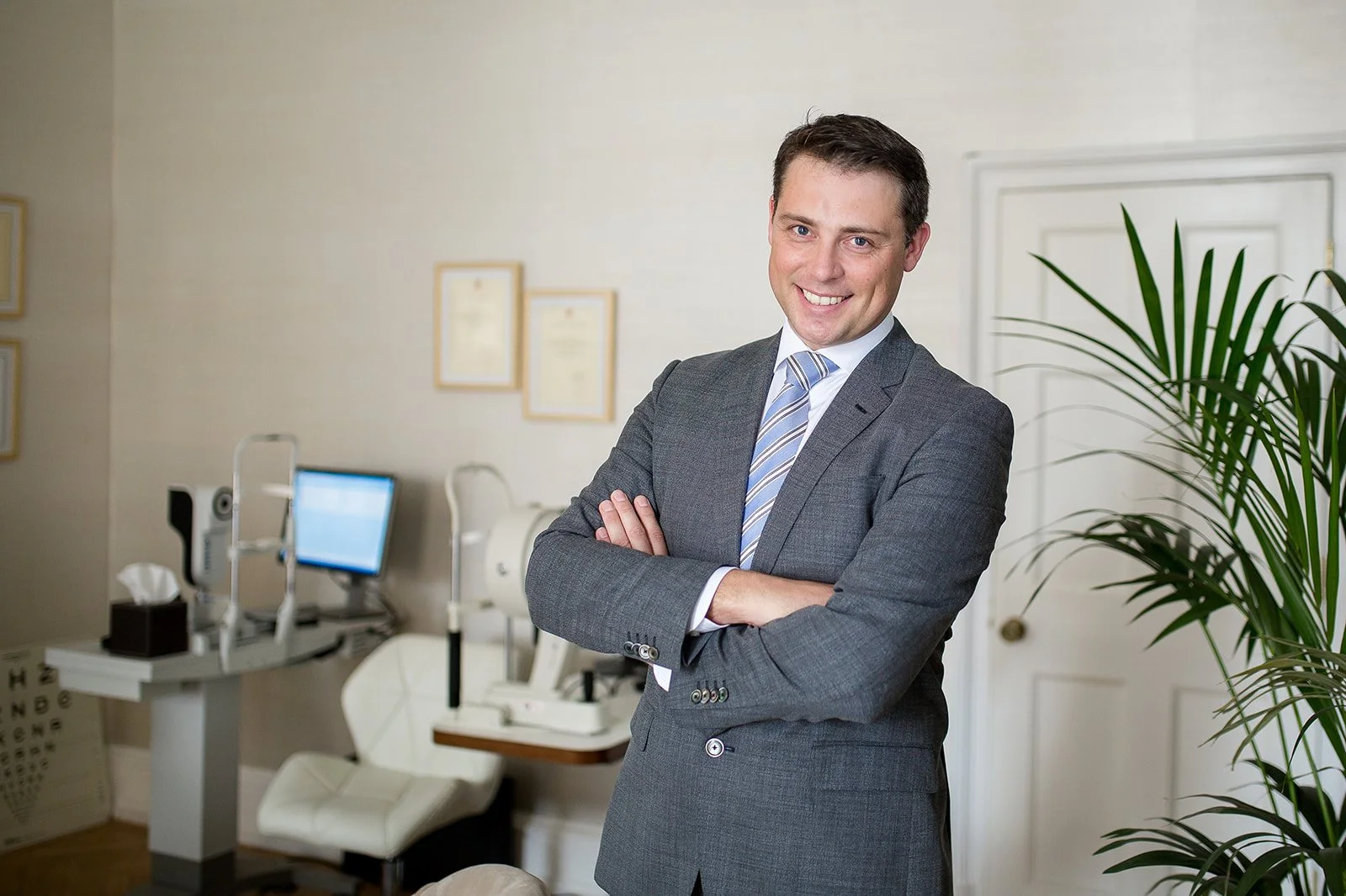 A man in a gray suit with a striped tie smiling and standing with arms crossed in an office setting with a potted plant, medical equipment, and framed certificates on the wall.