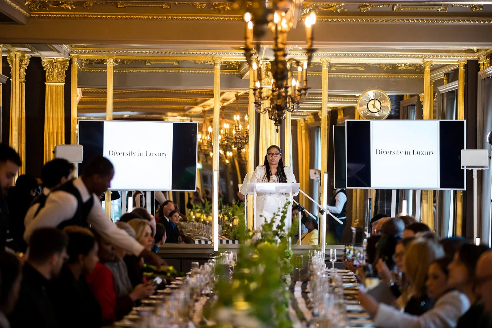 A woman speaking at a podium during a formal event in an opulent, gold-decorated room with chandeliers and large mirrors, with two large screens displaying the phrase "Diversity in Luxury," and an audience seated at a long banquet table.