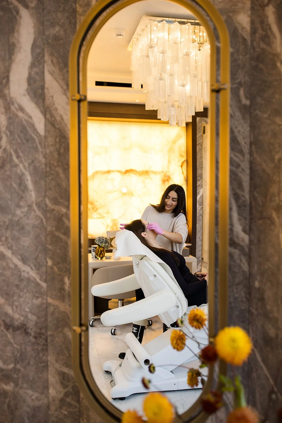 Reflection of a woman getting her makeup done at a beauty salon, seen through a mirror with a yellow frame, with decorative flowers at the bottom.