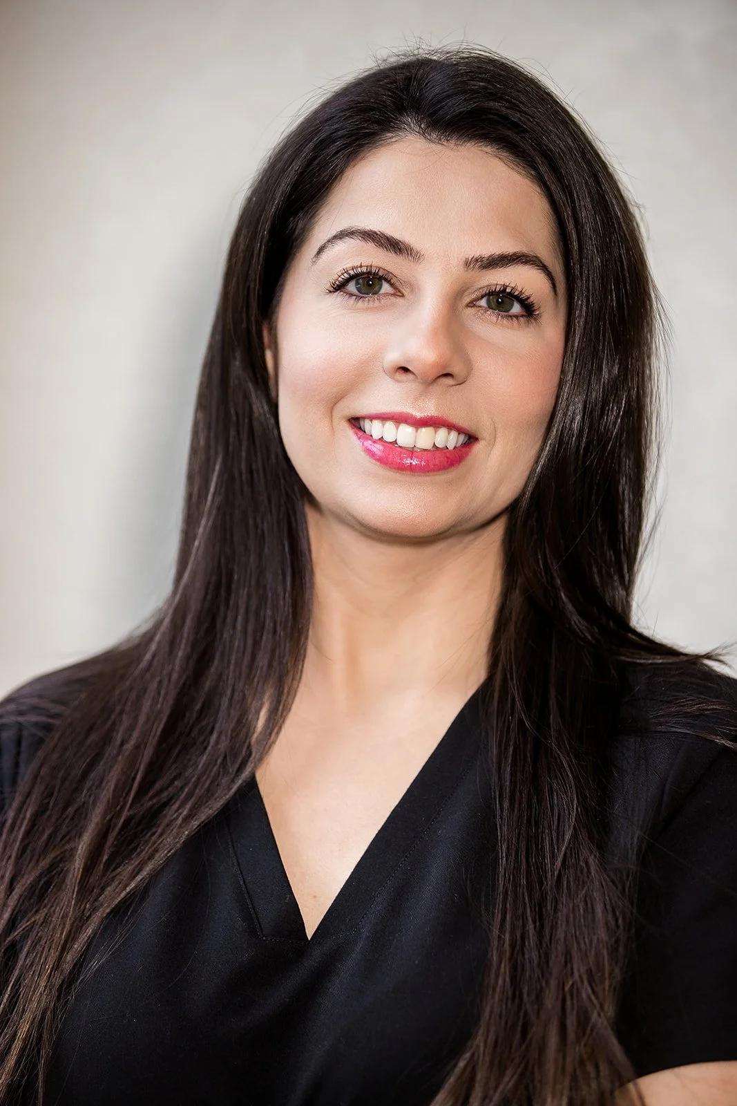 A woman with long dark hair, wearing a black top, smiling with her teeth showing, against a light-colored background.