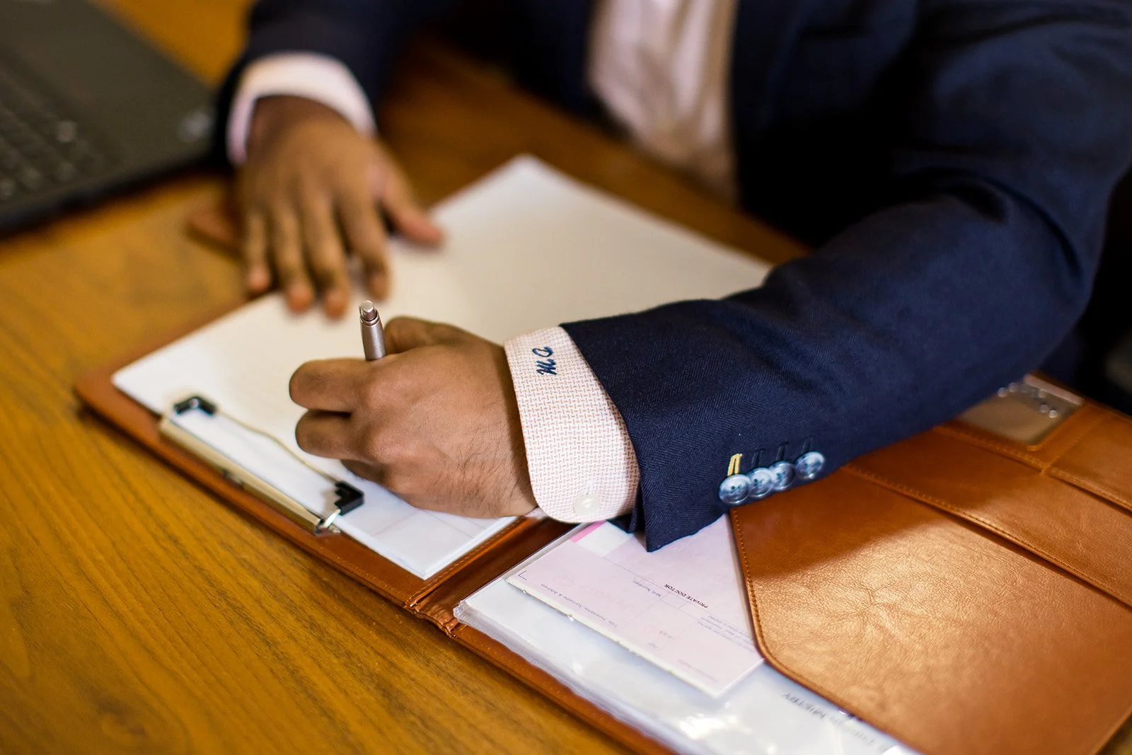 A person in a dark blue suit sits at a wooden desk, writing on a notepad with a silver pen. The person has a white shirt with embroidered initials 'M.G.' on the cuff. An open clipboard with papers is in front of them, and a laptop is partially visibl