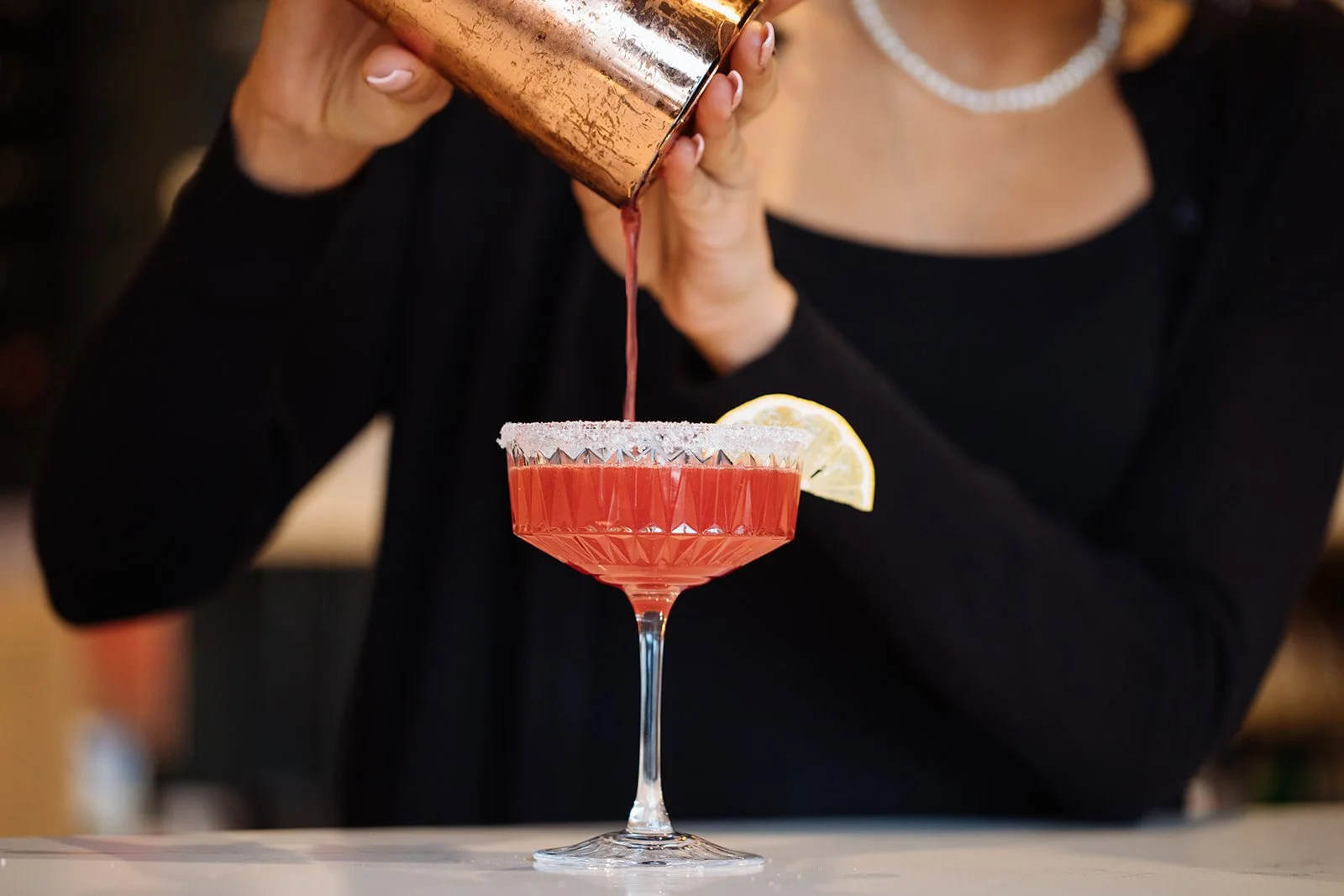 A person in black clothing is pouring a red cocktail from a copper shaker into a crystal glass rimmed with salt. The drink is garnished with a lemon wedge.
