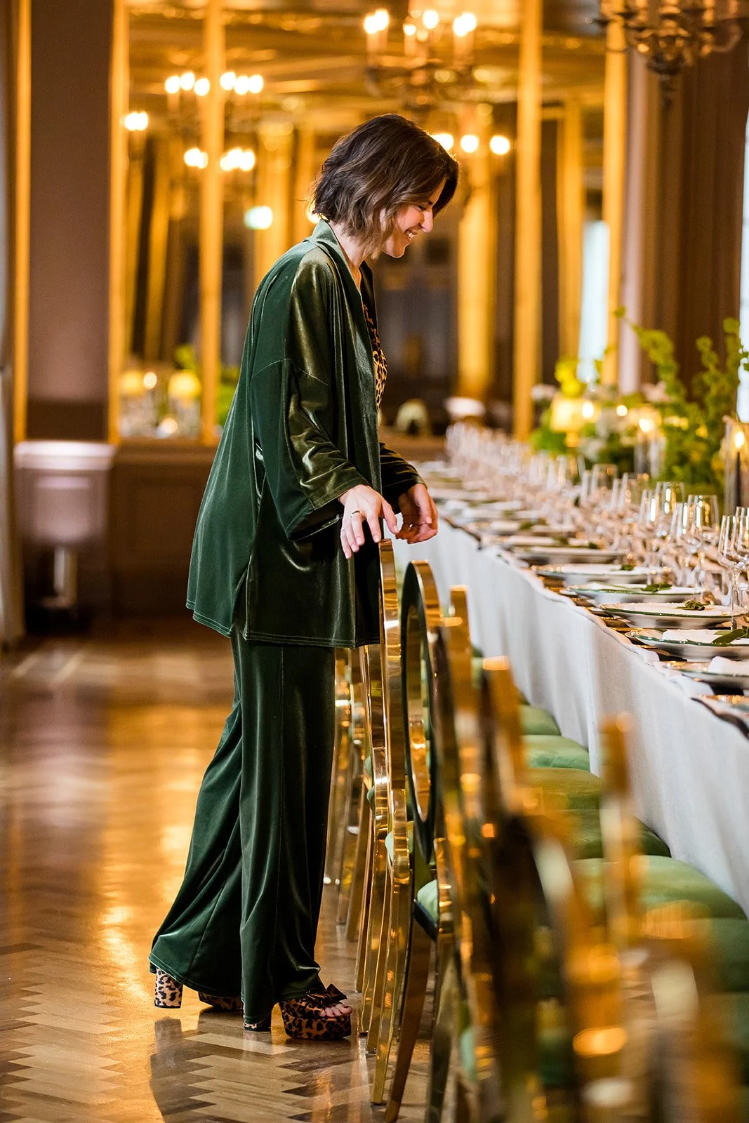 A woman standing by a long, elegantly set dining table in a luxurious room, looking down with a smile, dressed in a shiny green velvet jacket and matching pants, wearing leopard print high heels.