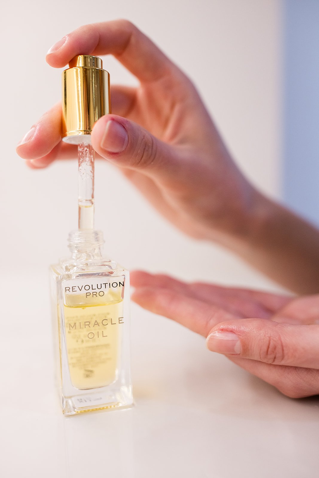 Person holding a gold-capped glass dropper bottle labeled "Revolution Pro Miracle Oil" dispensing oil into a small glass container.