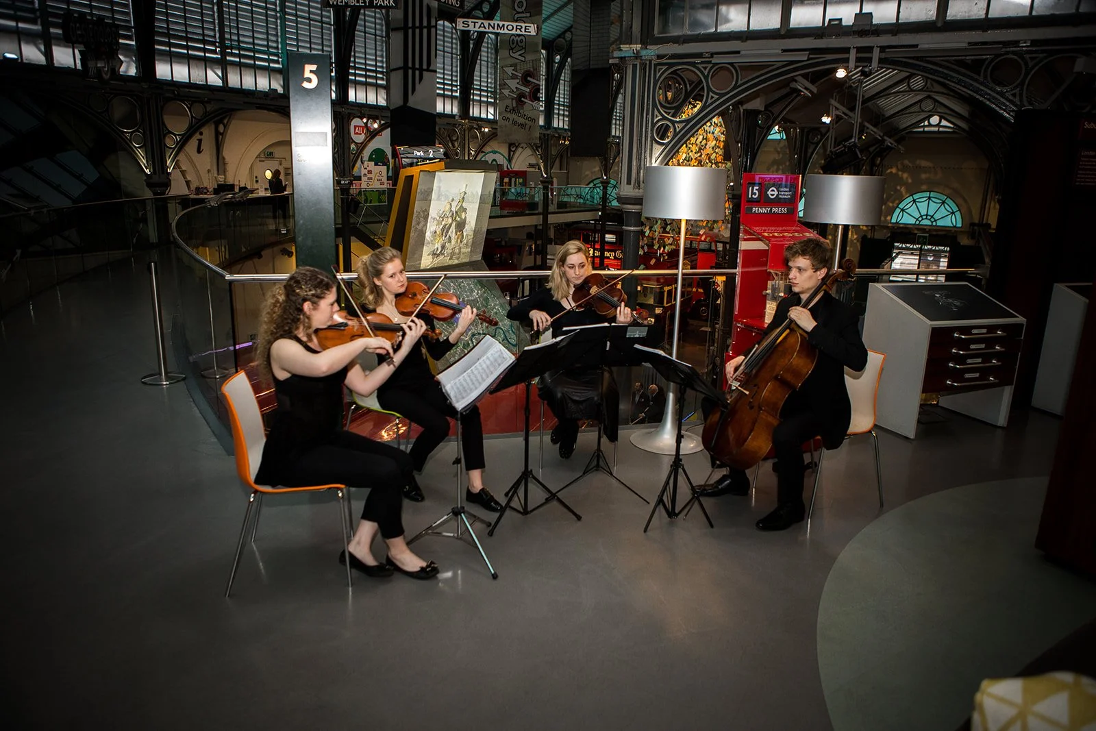 A group of four musicians performing with string instruments in an indoor space that resembles a shopping mall or public venue, with architectural details and signage visible in the background.