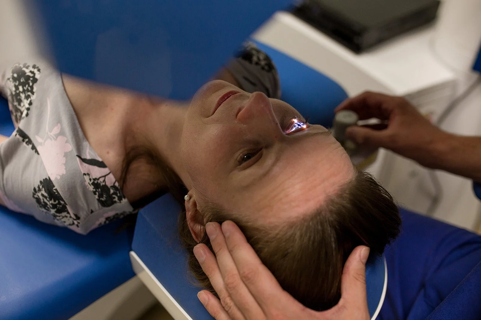 Woman lying on medical examination table with her eyes open, undergoing an eye exam with a doctor using a bright light. The woman has short brown hair and is wearing pearl earrings and a patterned blouse.