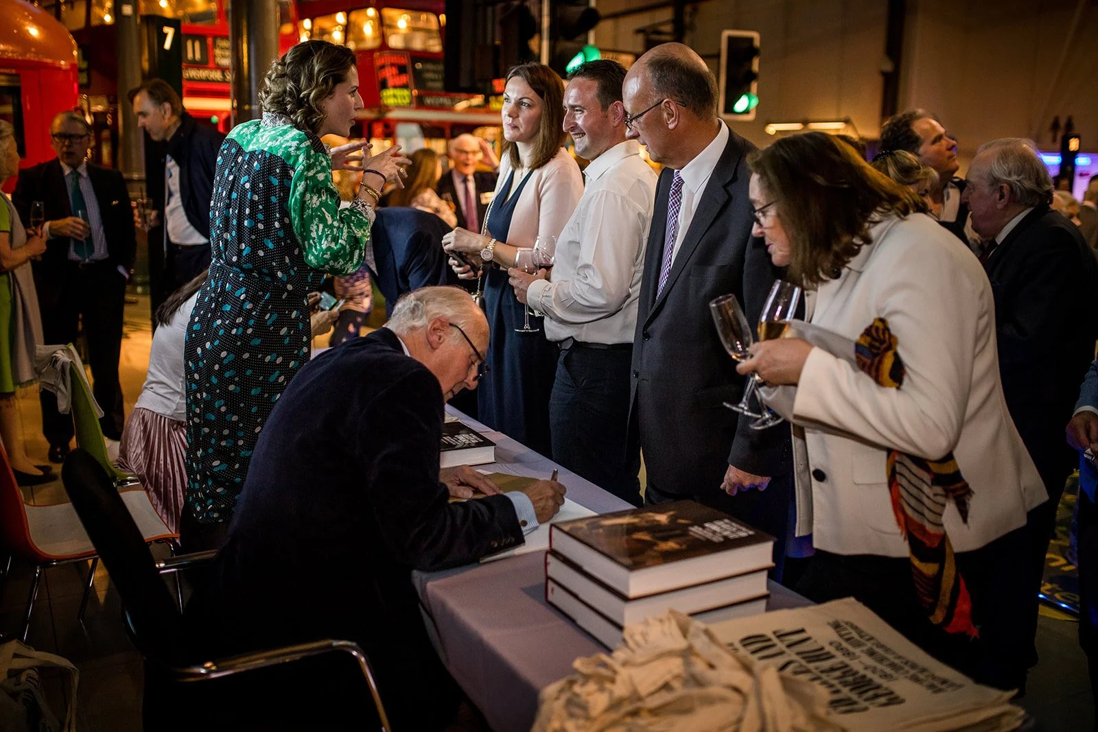 People gathered at a book signing event, with an author signing a book at a table, and others waiting or engaging in conversation, in a lively indoor setting with colorful lights.