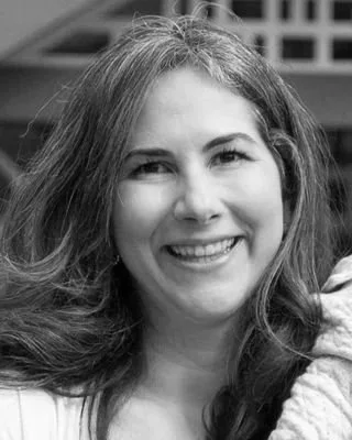 A smiling woman with shoulder-length hair, wearing a casual top, outdoors with a blurred background.