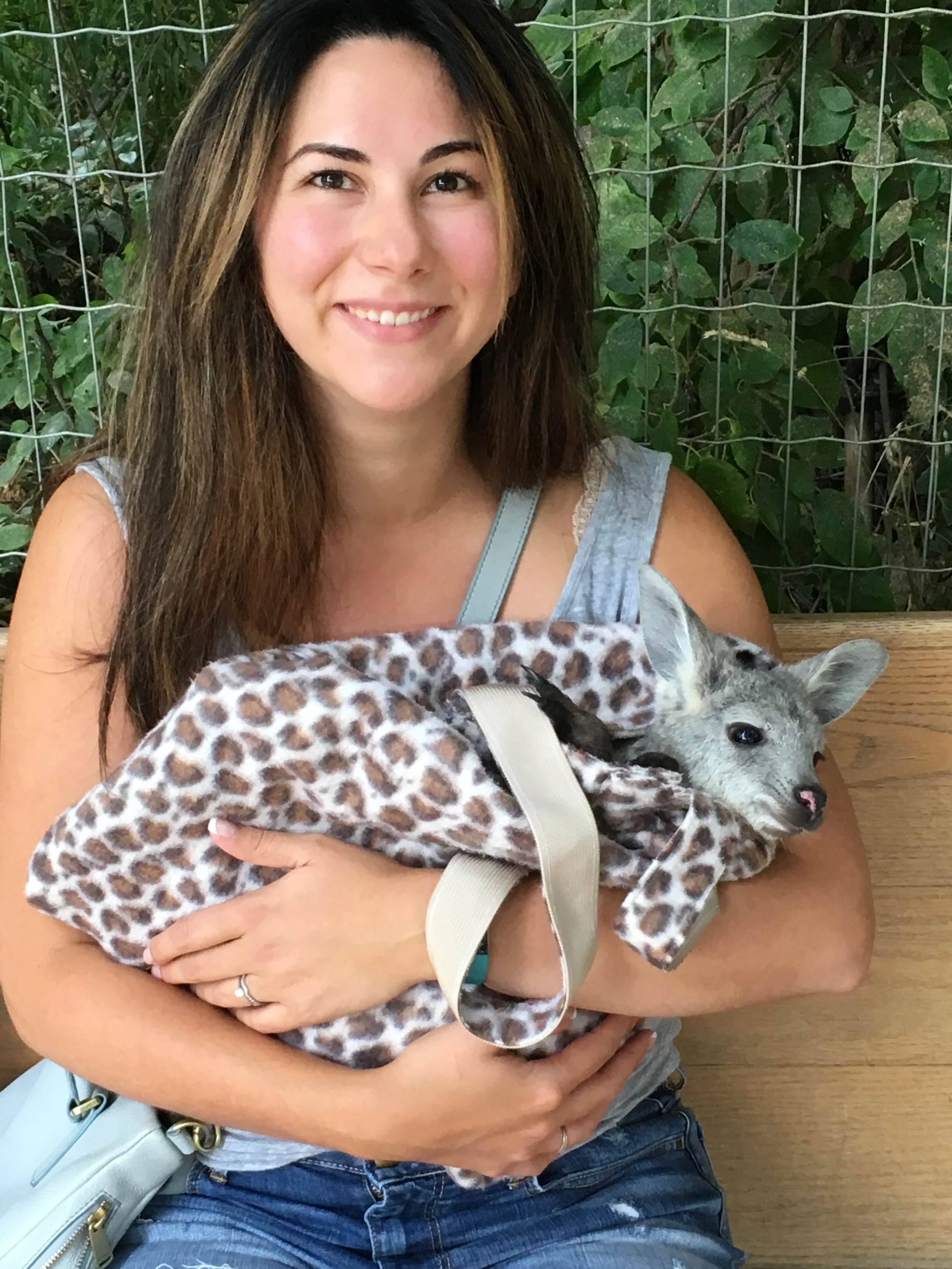 A woman with long brown hair is smiling and sitting outdoors on a wooden bench, holding a small young kangaroo wrapped in a leopard-print blanket.