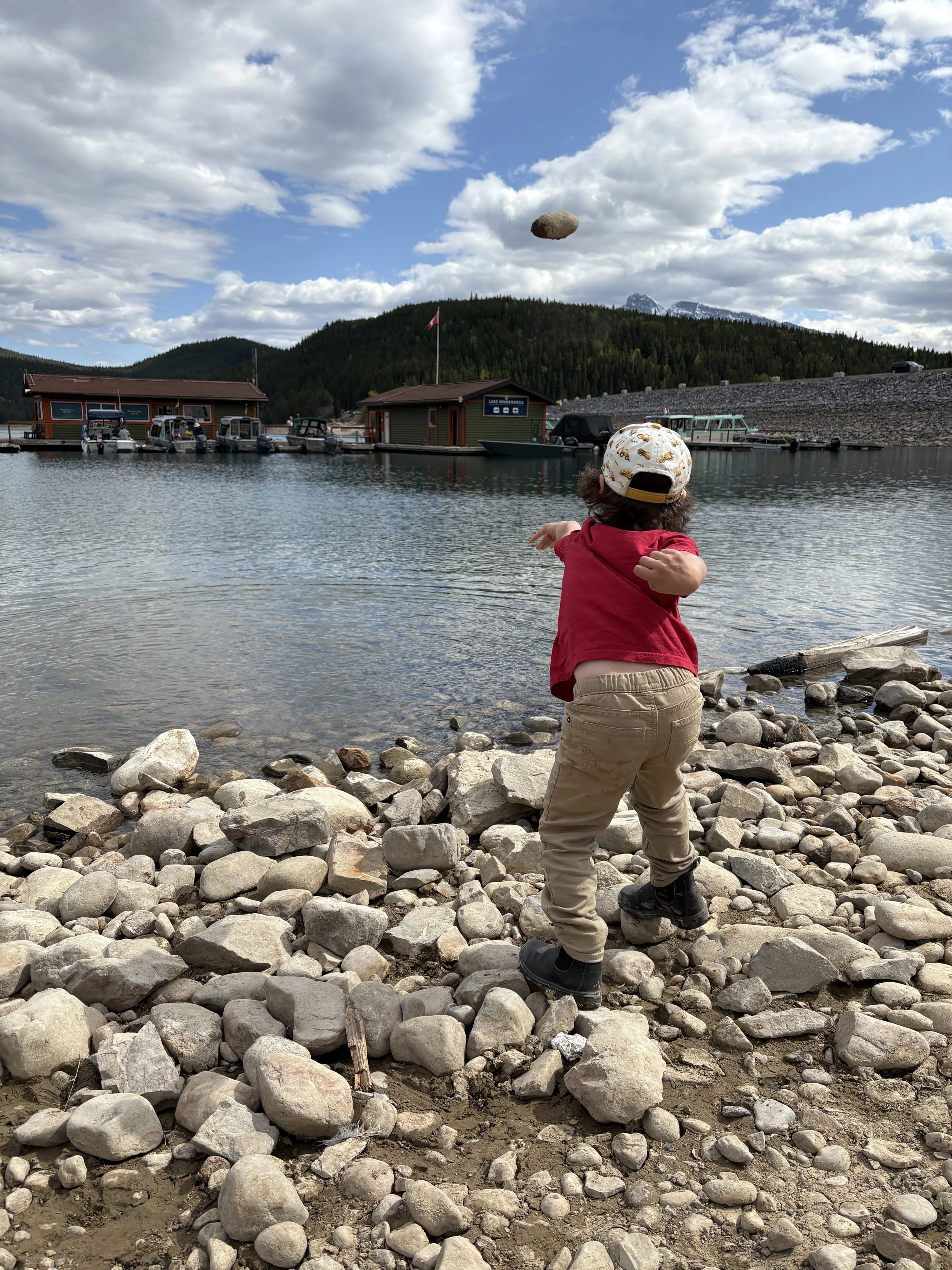 A young child wearing a white cap, red shirt, beige pants, and black shoes is throwing a rock into a lake on a rocky lakeshore. The scene includes boats docked at a marina and green mountains under a partly cloudy sky.