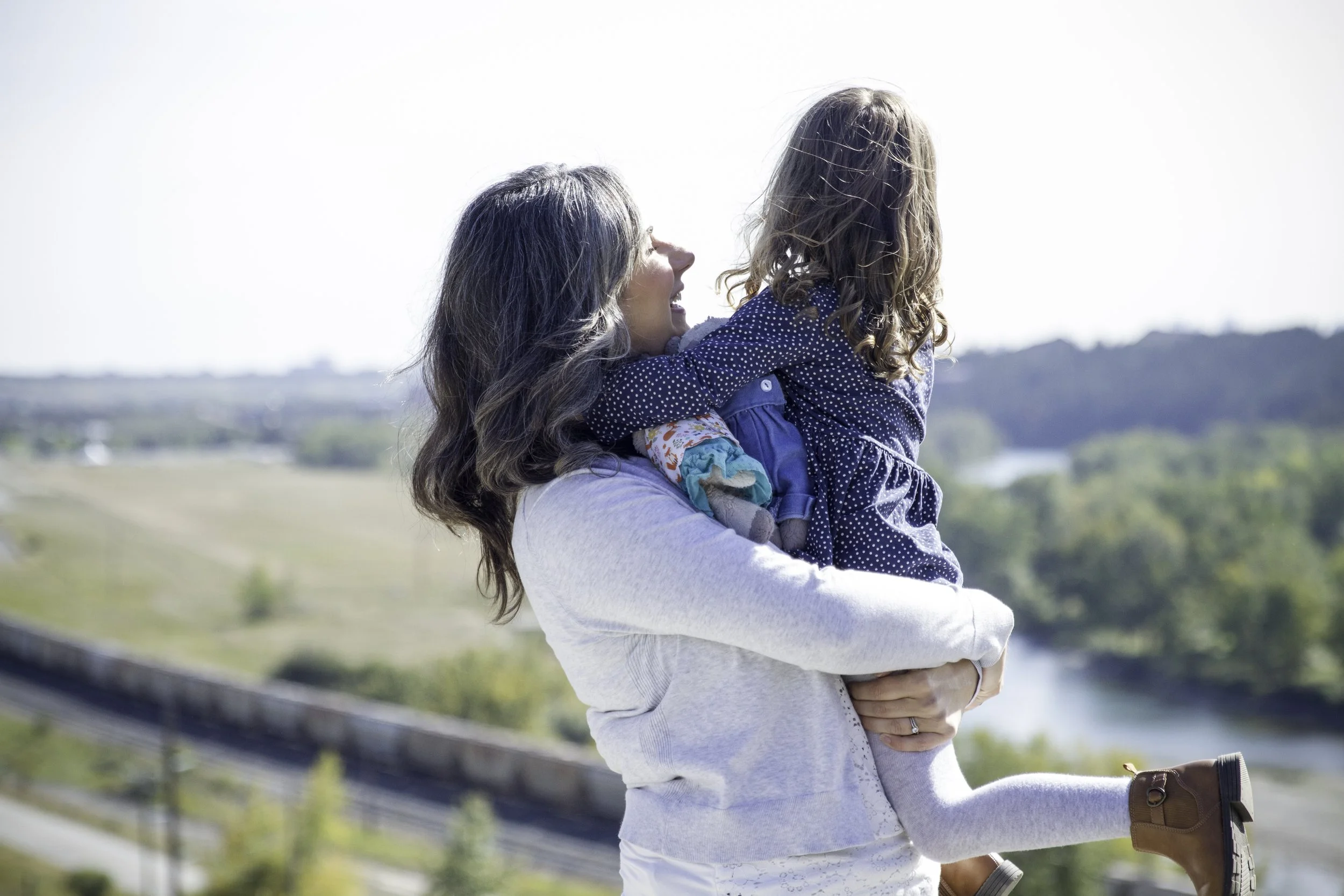 A woman holding a young girl outdoors, with her arms around her and the girl smiling. The background features a scenic landscape with a river, trees, and hills under a clear sky.
